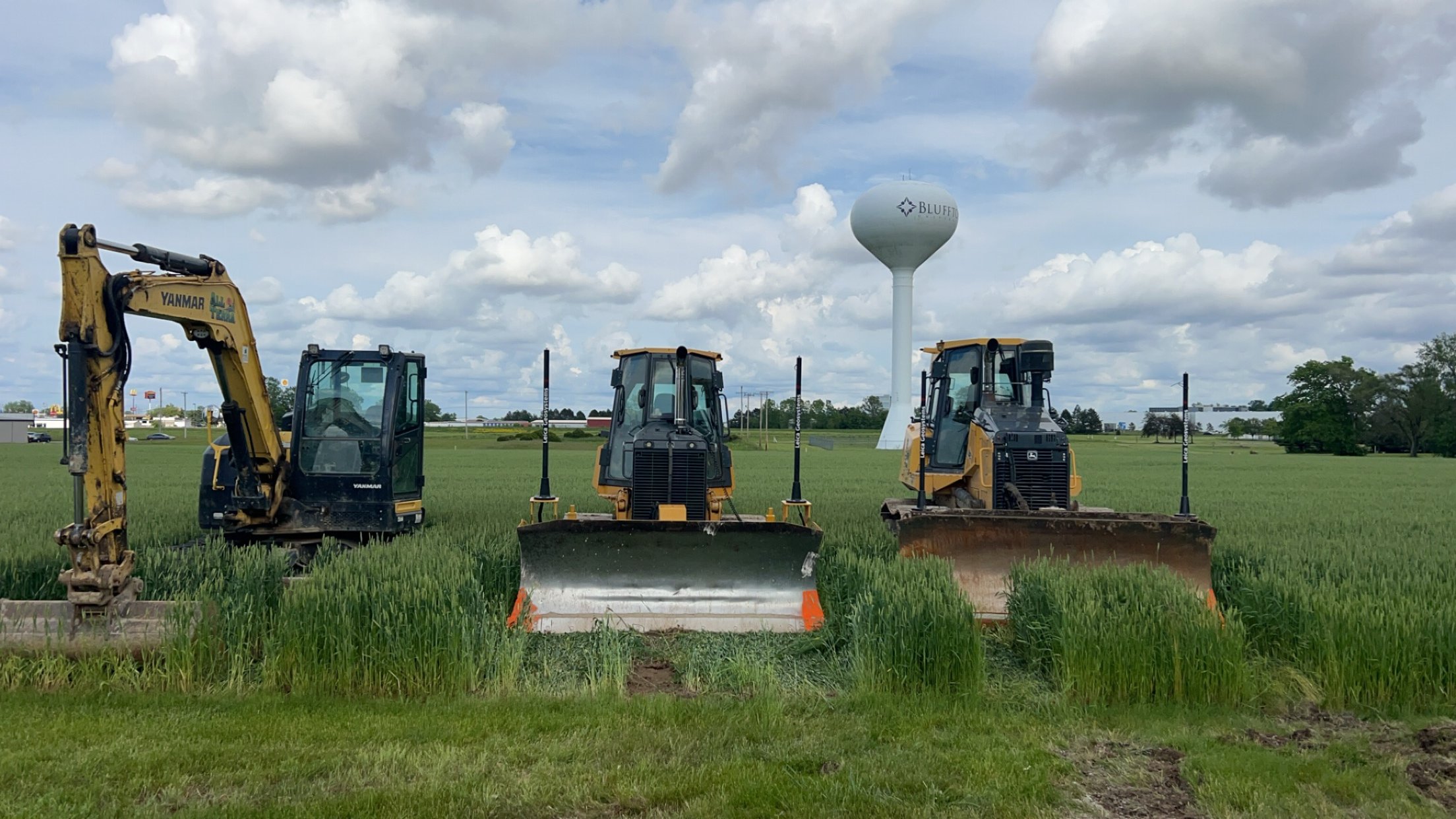 Three construction vehicles, a backhoe, a compact excavator, and a bulldozer, are lined up in a field of green grass under a partly cloudy sky. In the background, there is a water tower with the word 'BLUFF' written on it, and some distant buildings and trees.