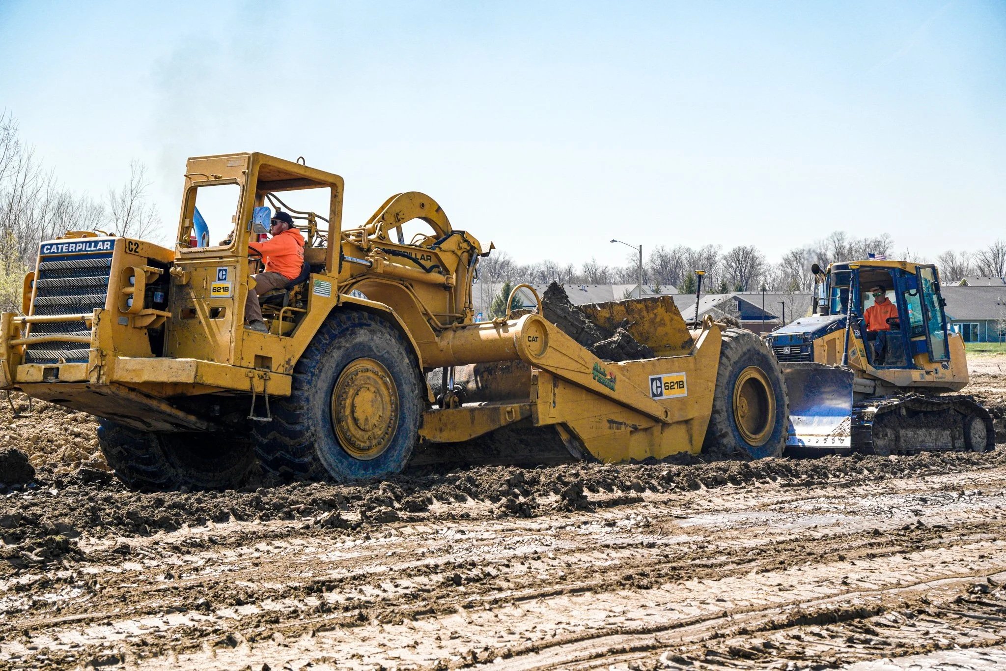 Two construction workers operating yellow bulldozers on a dirt site with houses and leafless trees in the background.