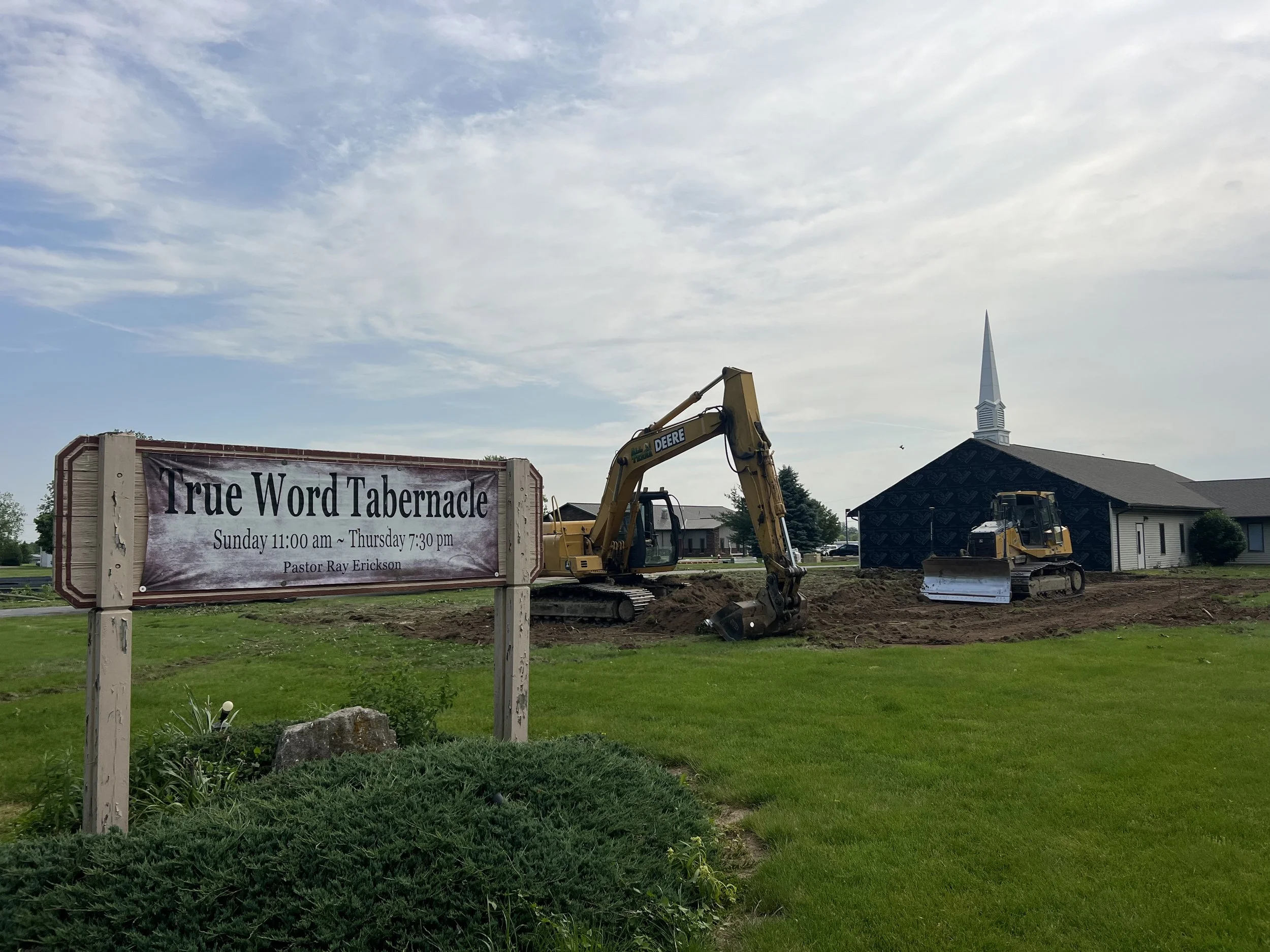 Construction site with two mini excavators and a church steeple in the background. A sign reads "True Word Tabernacle" with service times and pastor's name.