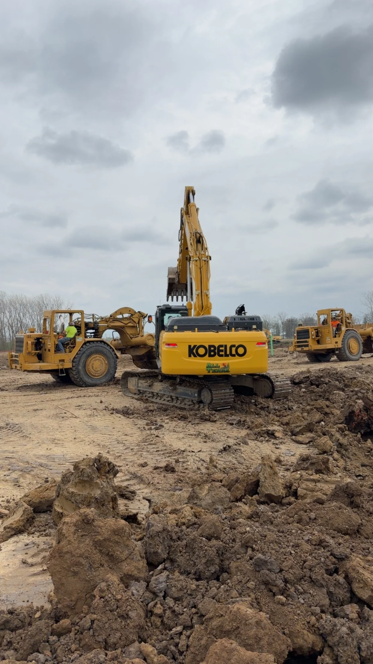 Construction site with a yellow excavator labeled 'Kobelco' digging into the ground, two workers in safety gear operating nearby, and a muddy landscape under a cloudy sky.