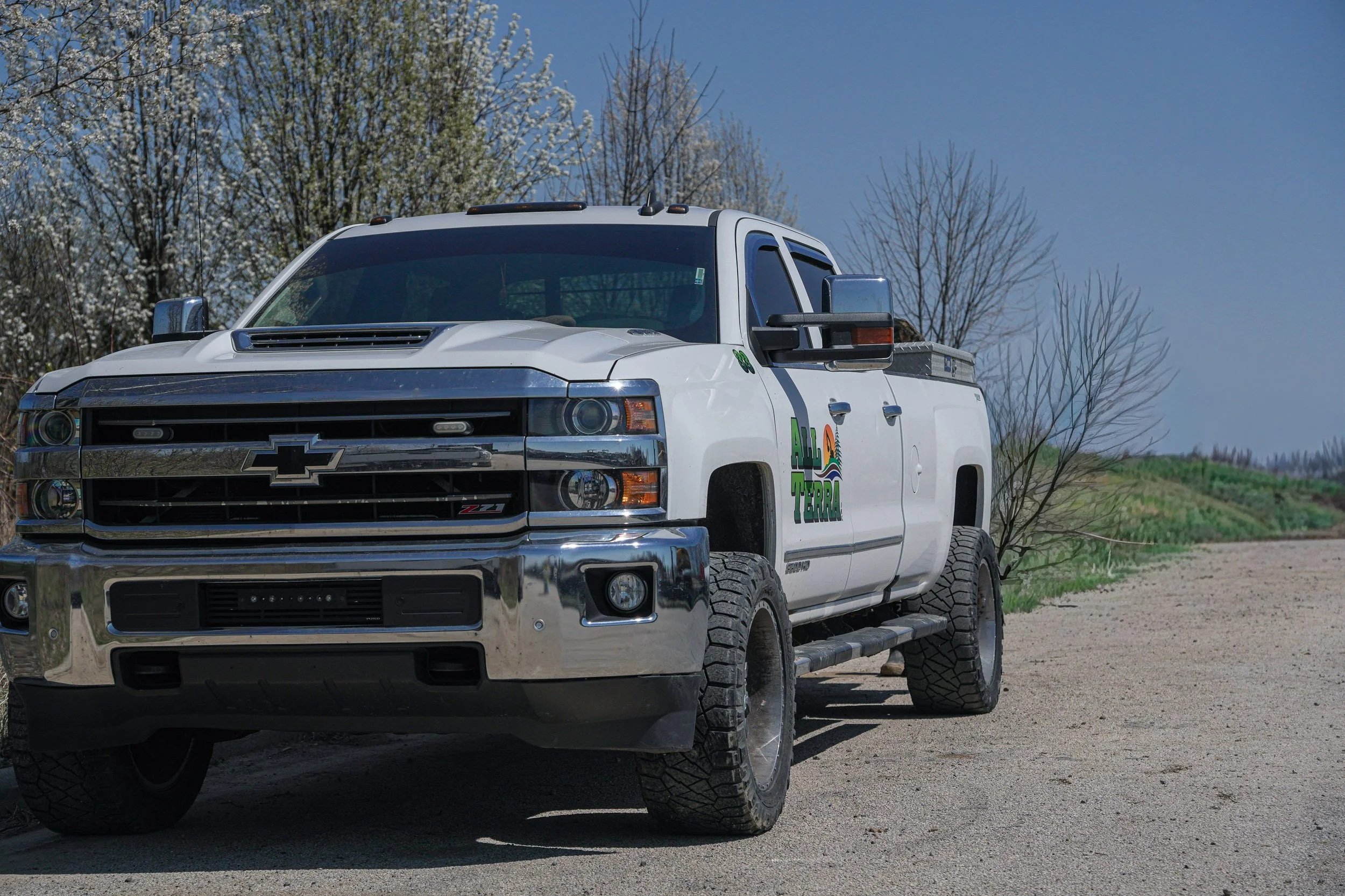 White Chevrolet pickup truck with a logo that reads 'Agro Tierra' on the door, parked on a dirt road under a blue sky. Trees with budding leaves are in the background.
