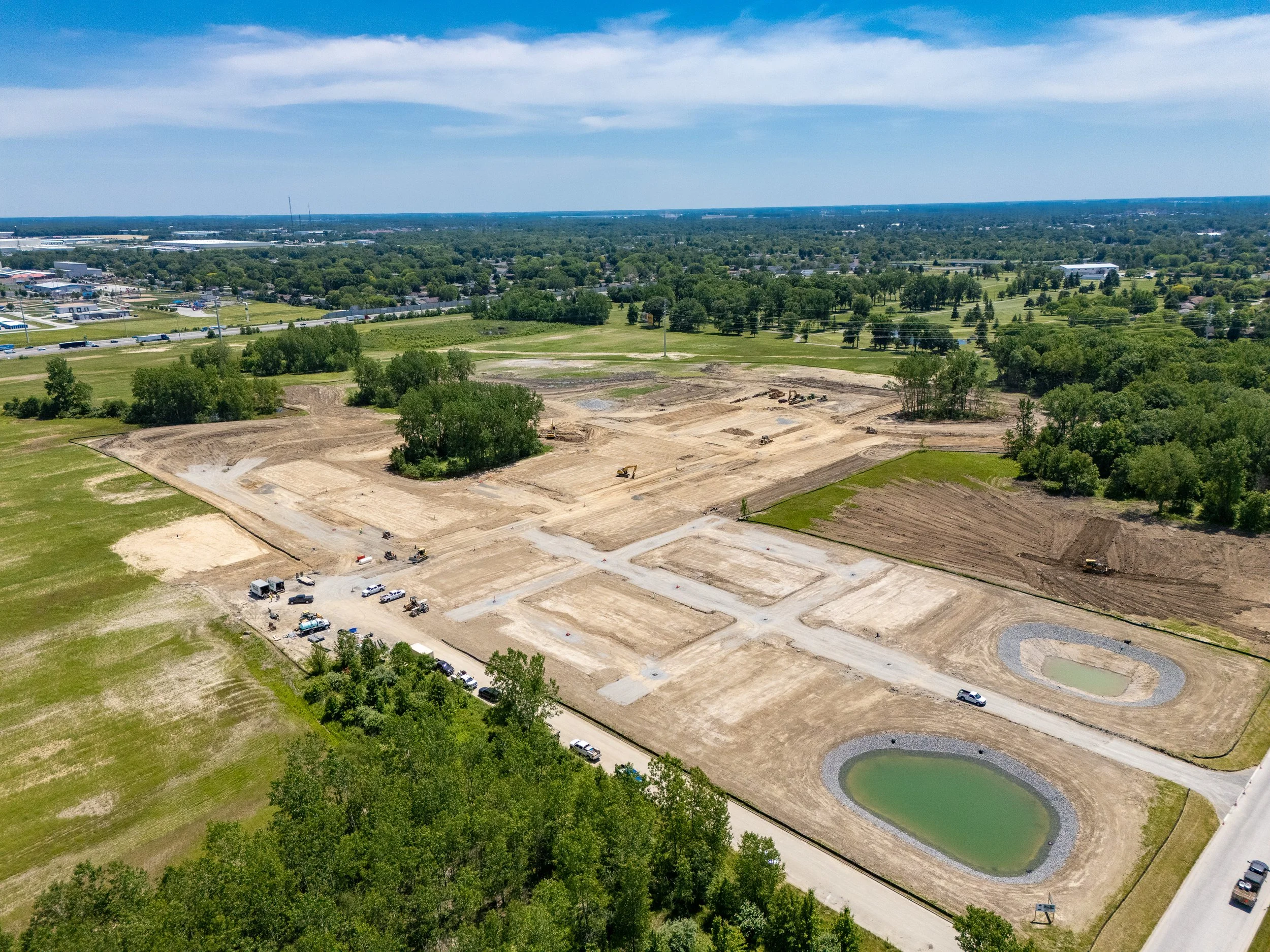 Aerial view of a construction site next to a pond, with green trees, a pond, and a commercial area in the distance.