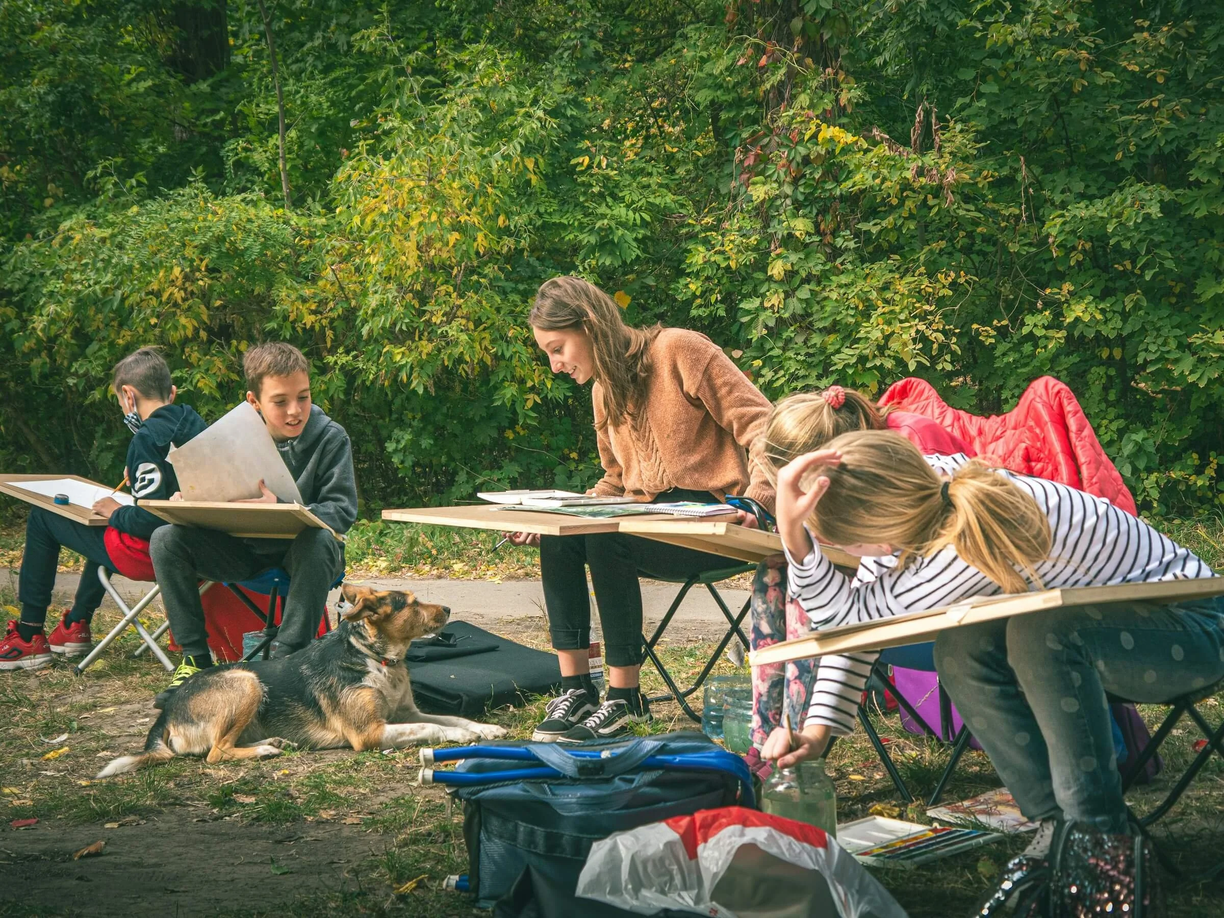 Children and a woman sitting and lying on benches outdoors with trees in the background. There are art supplies, papers, water bottles, and a dog on the ground.