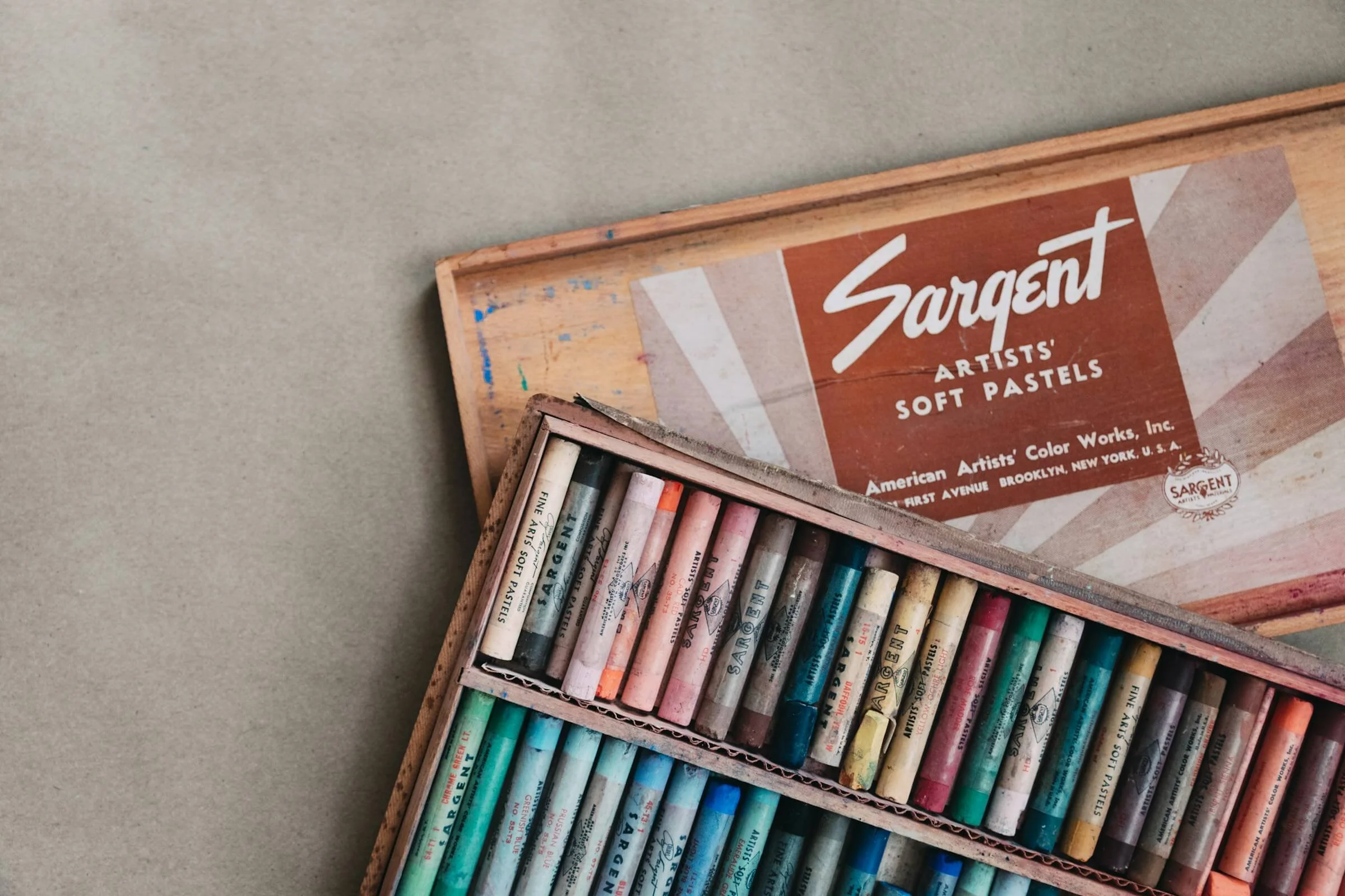 Part of a wooden art supplies box with pastel sticks and a vintage Sargent Artists' Soft Pastels box lid.