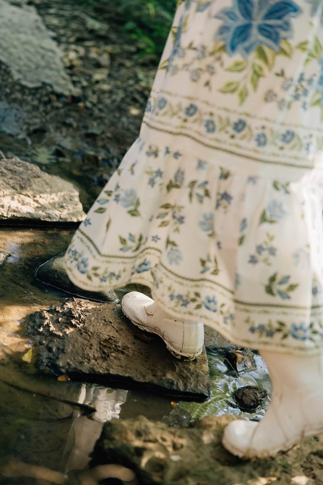 Close-up of a person's leg in a floral dress and white boot stepping on rocks in a shallow stream.
