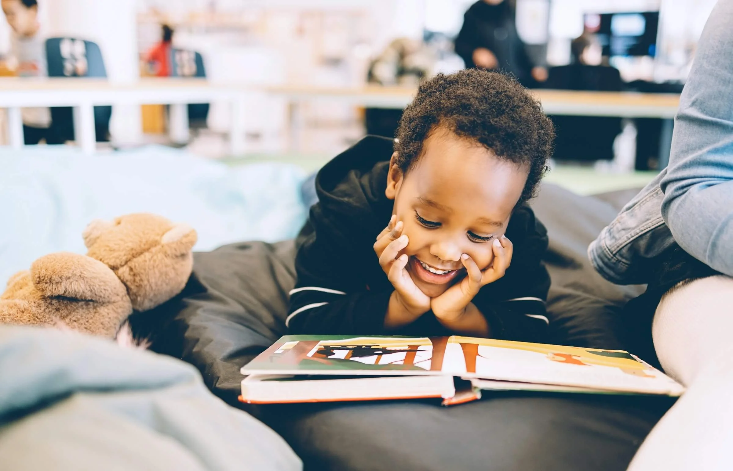 A young child with curly hair smiling and resting their face in their hands, lying on their stomach in front of a colorful book, with a teddy bear beside them, in a bright indoor setting.