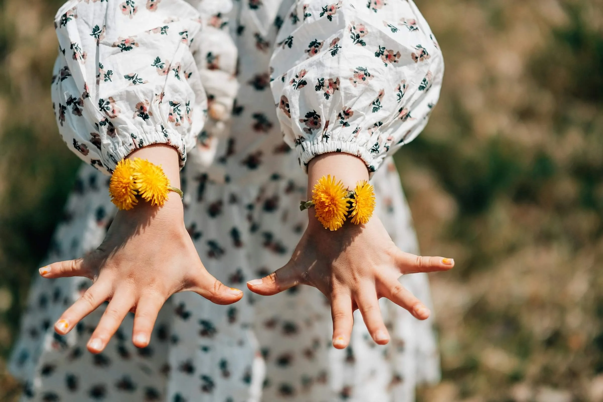 Person wearing a floral blouse holding yellow dandelions on their wrists, with multiple hands posed in various positions around them.