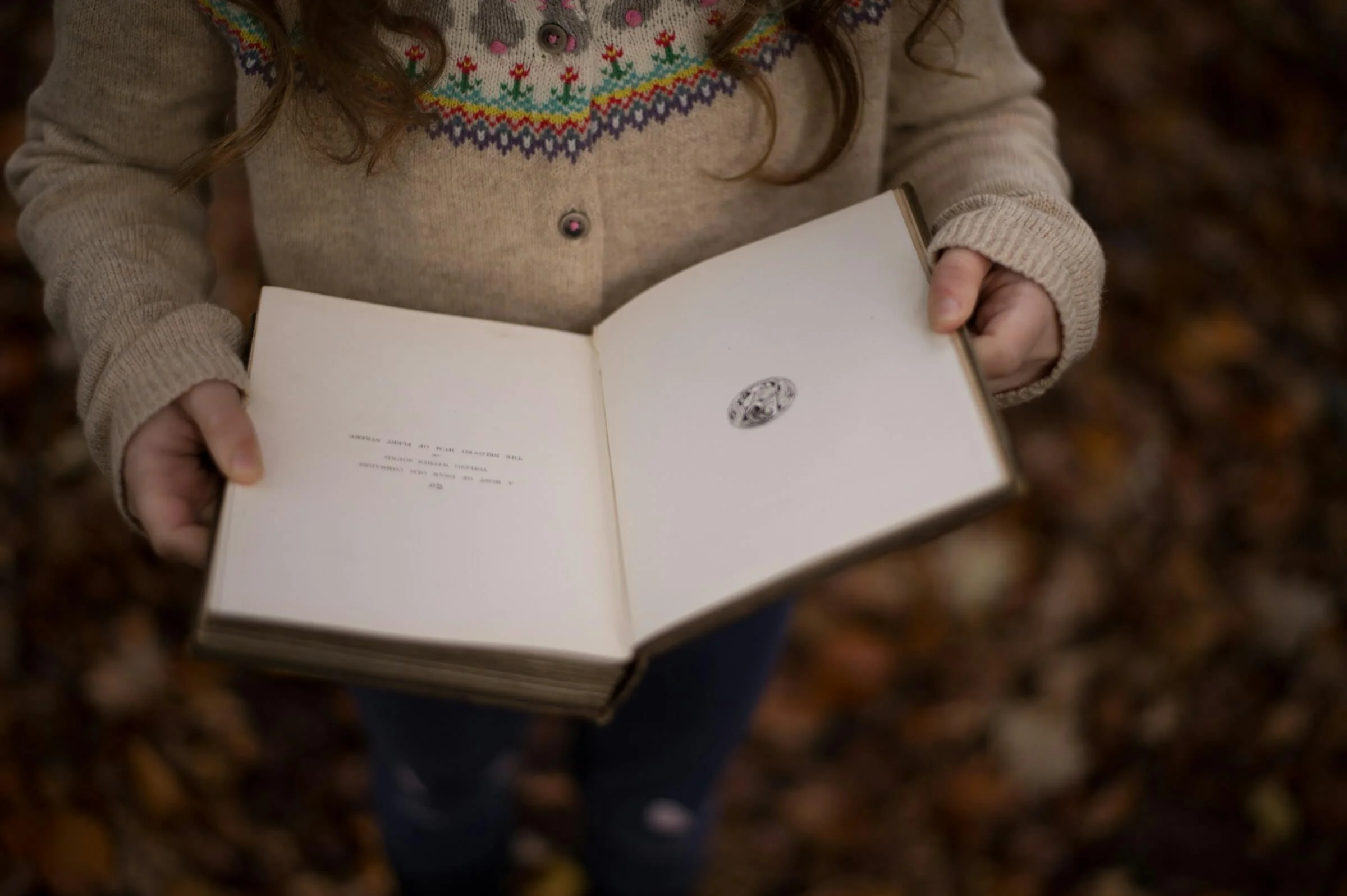 A person holding an open book with blank pages, wearing a beige sweater with colorful embroidery around the collar, standing on a bed of autumn leaves.