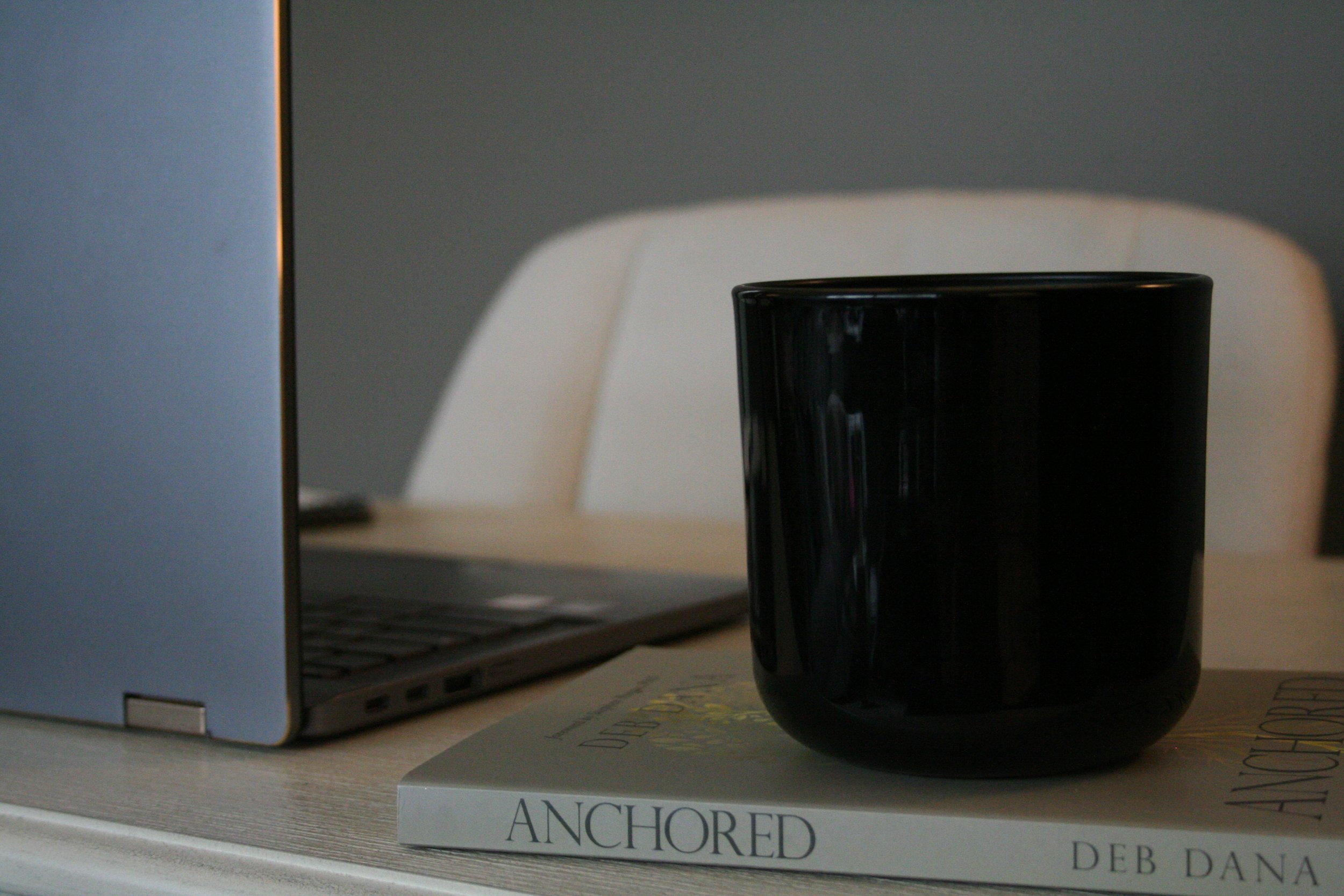 A black mug placed on a white book titled 'Anchored' resting on a desk next to a laptop.