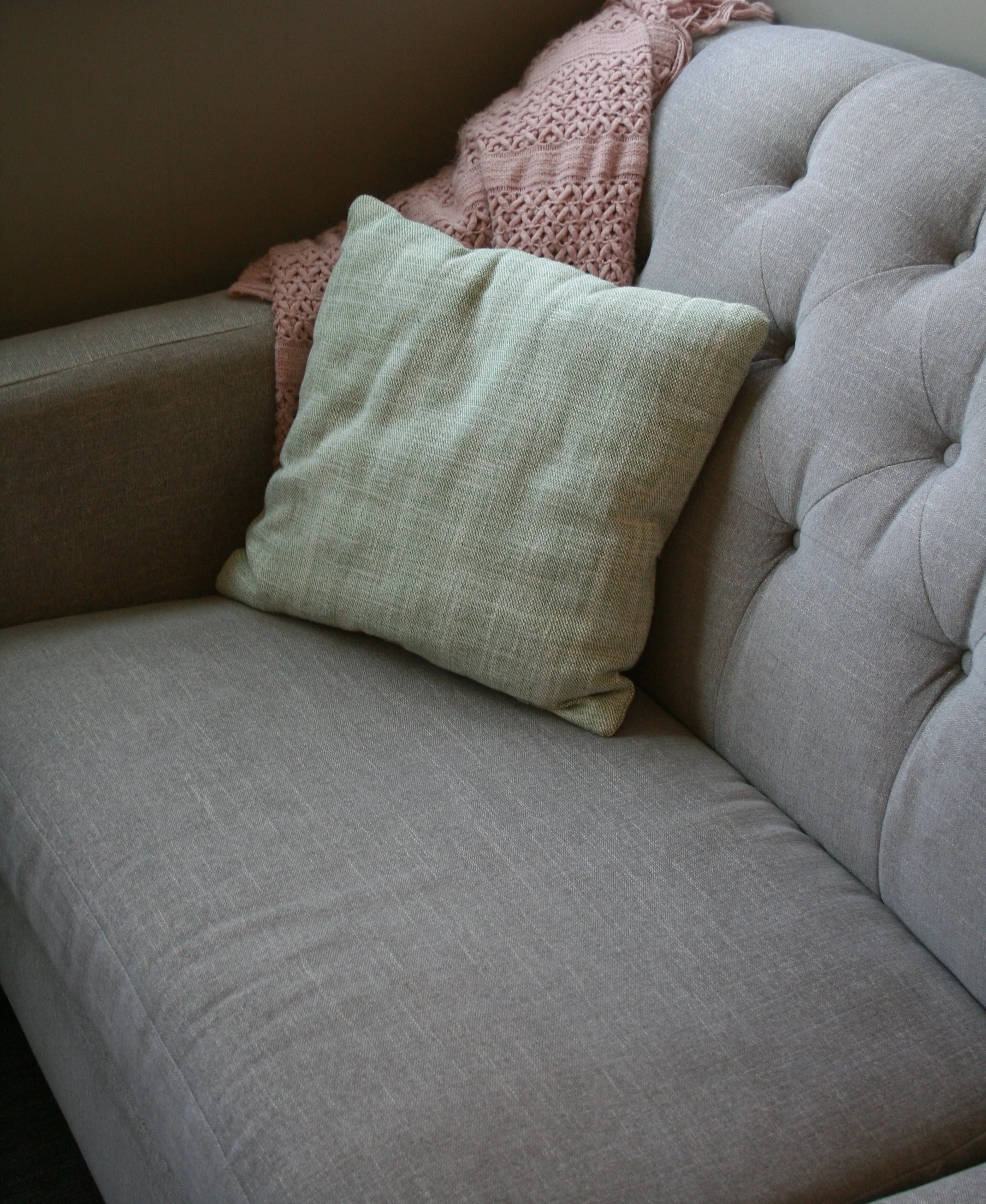 Close-up of a beige upholstered couch with a light green pillow and a pink knitted blanket draped over the back.