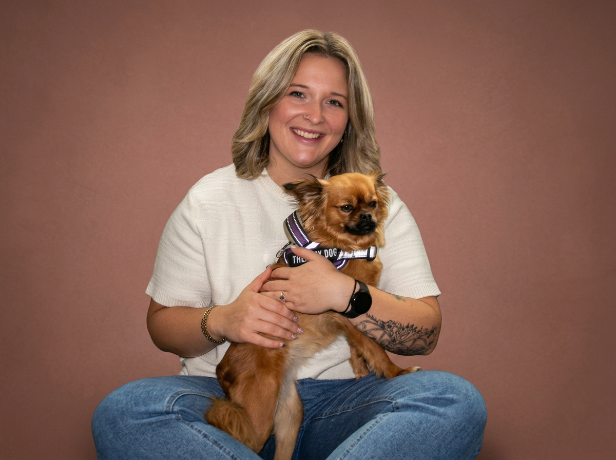 Smiling woman sitting cross-legged on a brown background, holding a small brown dog with a purple harness, with tattoos on her arm and wearing a watch.