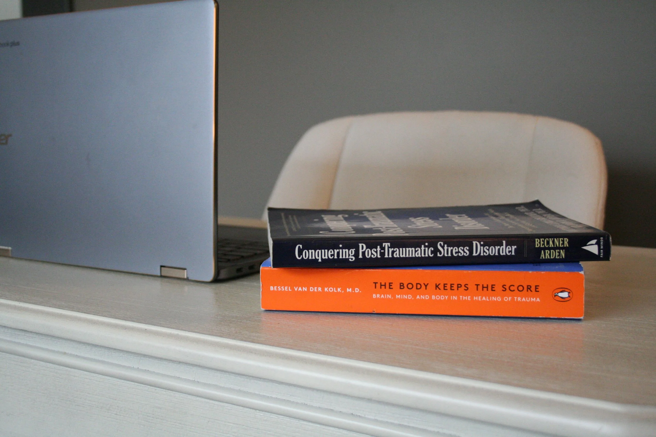 A stack of books on a desk next to a white chair, with one book titled 'Conquering Post-Traumatic Stress Disorder' and an orange book titled 'The Body Keeps the Score'. A silver laptop is partially visible on the left side of the image.