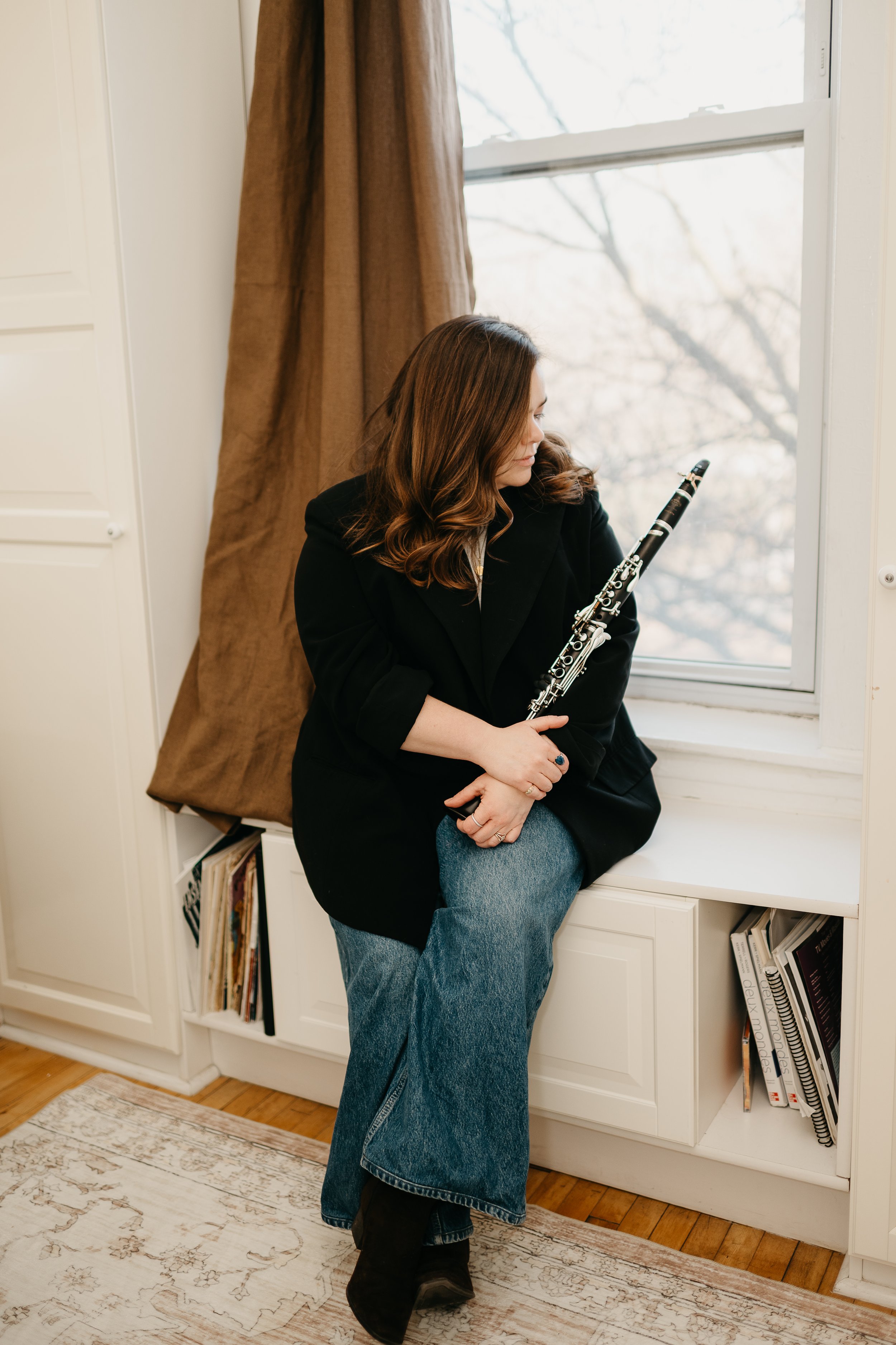 A woman with brown wavy hair sitting on a windowsill, holding a clarinet, wearing a black blazer, blue jeans, and black boots with a calm expression, next to a window with brown curtains and bookshelves below.