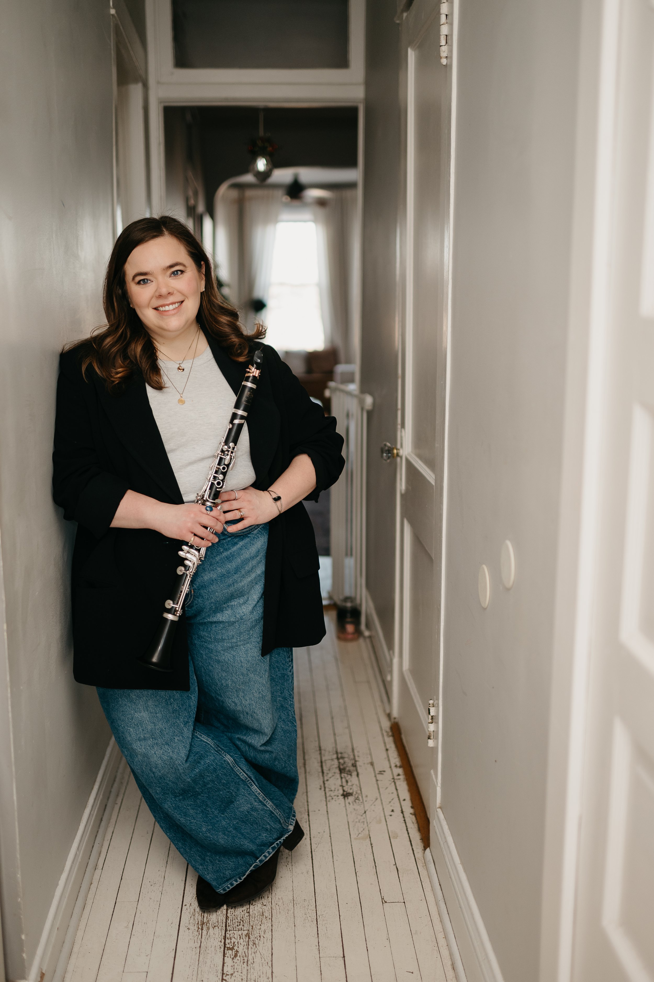 A woman with brown hair holding a clarinet stands in a narrow hallway with white walls and wooden flooring. She is smiling, wearing a black blazer over a light-colored shirt and blue jeans.