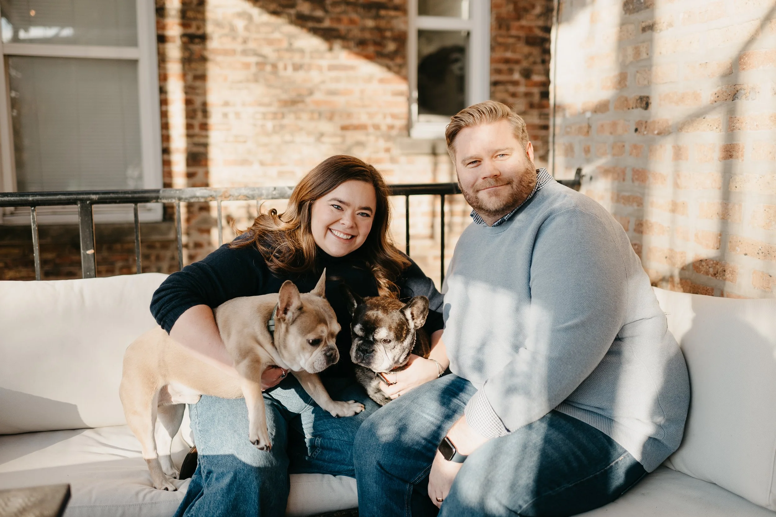 A smiling woman and man sitting on a white couch, each holding a bulldog puppy, in front of a brick wall and open window.