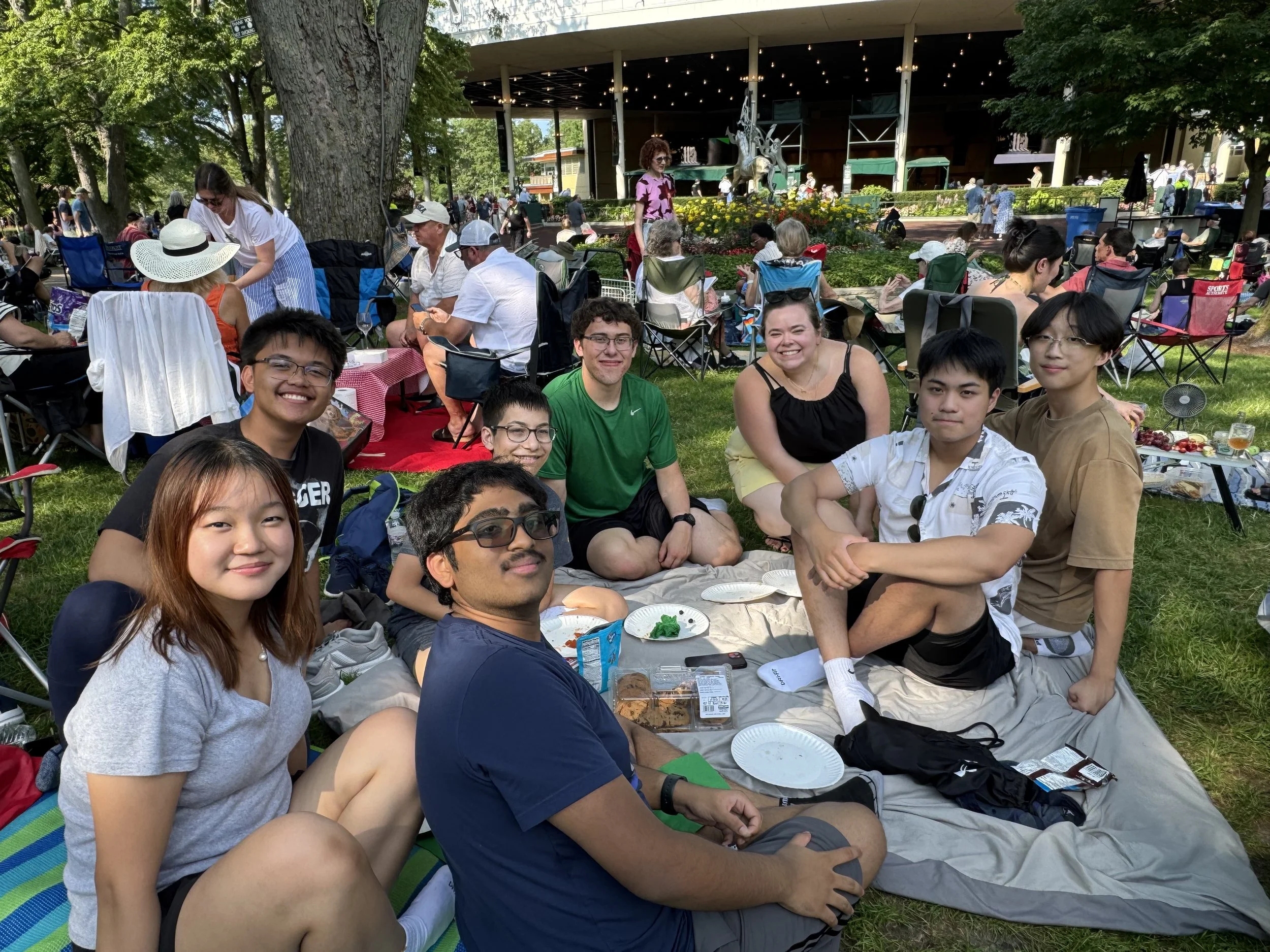 A group of people sitting on a blanket and lawn chairs during an outdoor gathering on a sunny day, with several people standing and walking in the background near a building and trees.