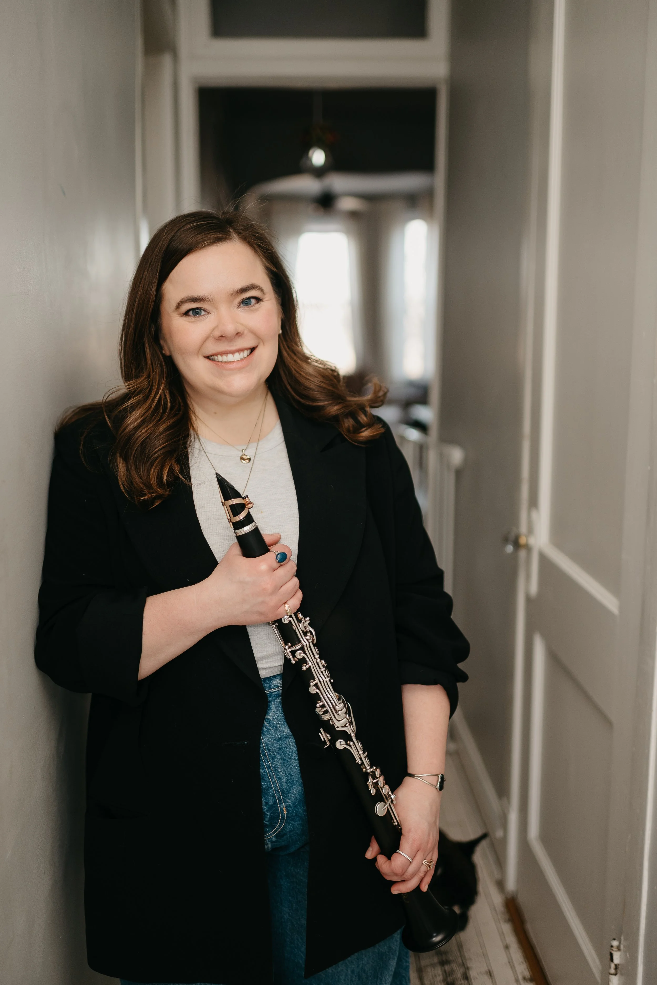 A woman with brown hair, wearing a black blazer, white shirt, and blue jeans, holding a clarinet and smiling while standing in a narrow hallway with gray walls and white doors.
