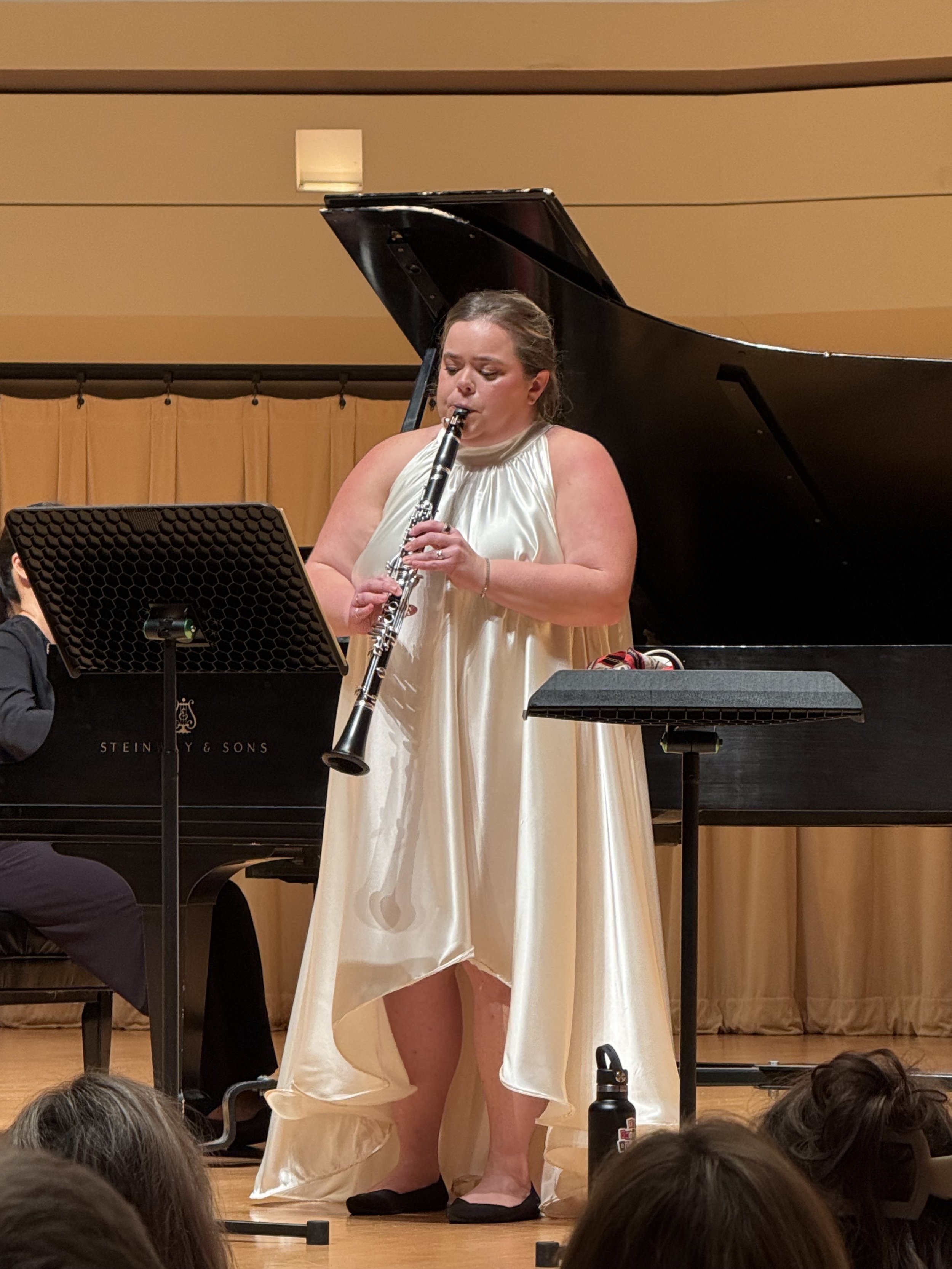 A woman in a cream-colored gown with a high slit is playing a clarinet on stage, with a grand piano and music stand in the background, and audience members in the foreground.