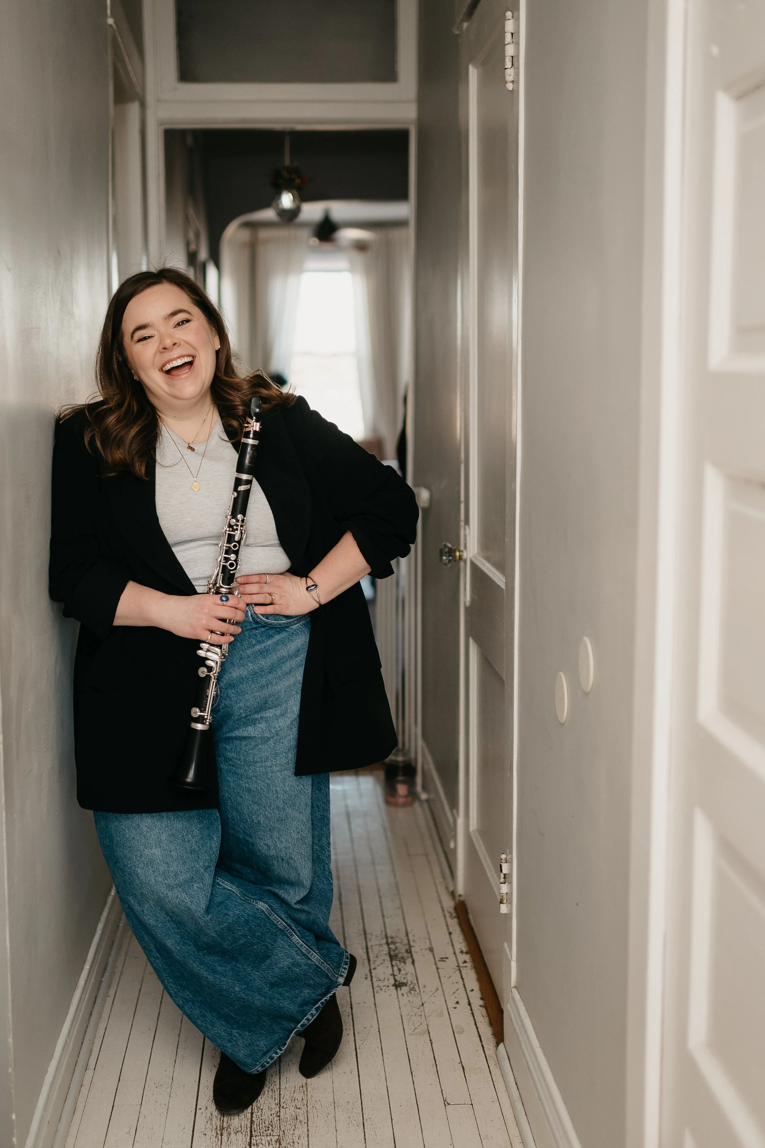 A woman with brown hair, wearing a Black blazer, gray t-shirt, and wide blue jeans, stands in a narrow hallway with wood flooring. She is holding a clarinet and smiling at the camera.