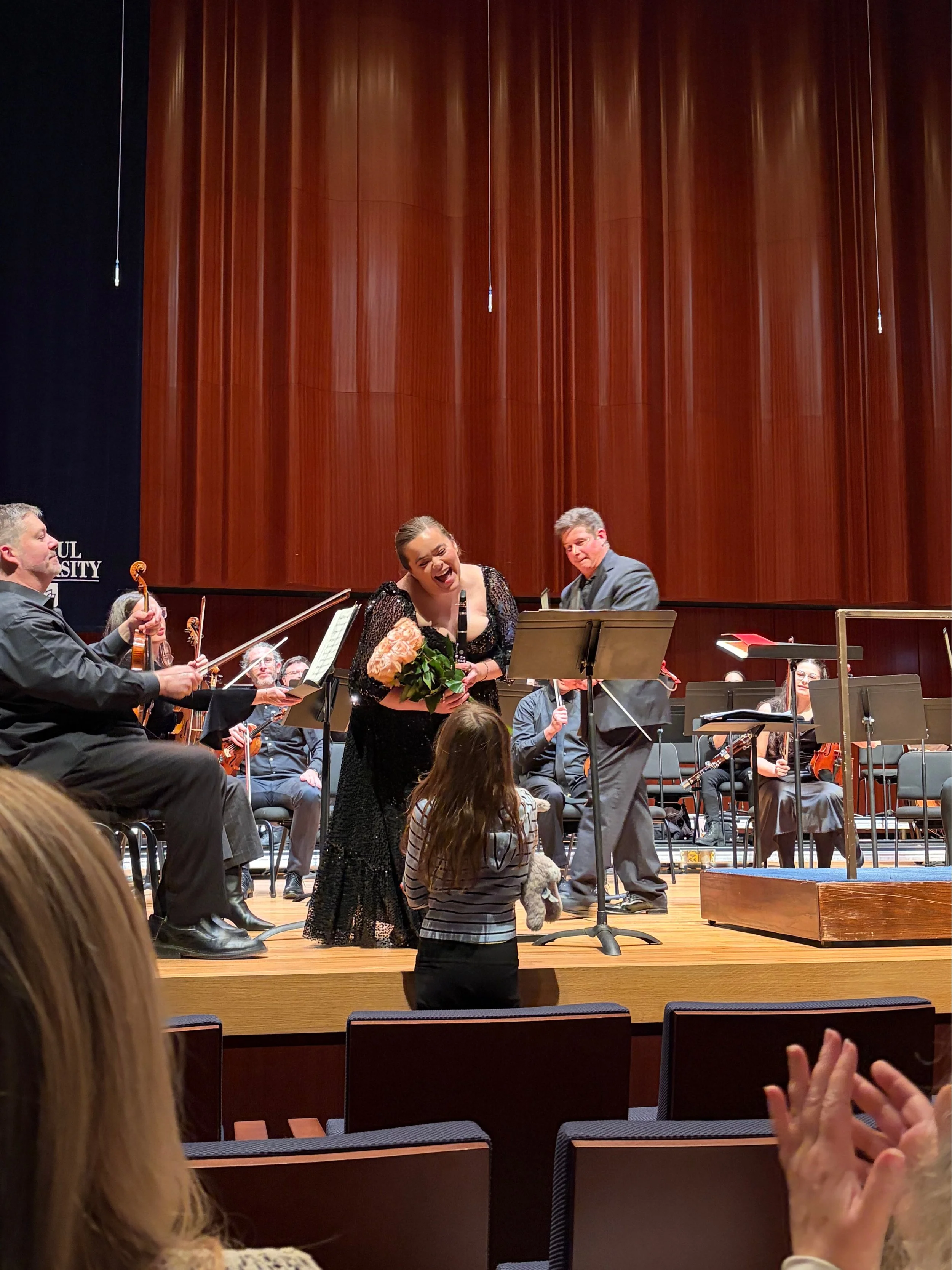 A live orchestral performance on stage at a concert hall, with a woman in a black dress holding flowers and smiling, surrounded by orchestra members including a violinist and conductor. Audience members are visible in the foreground.