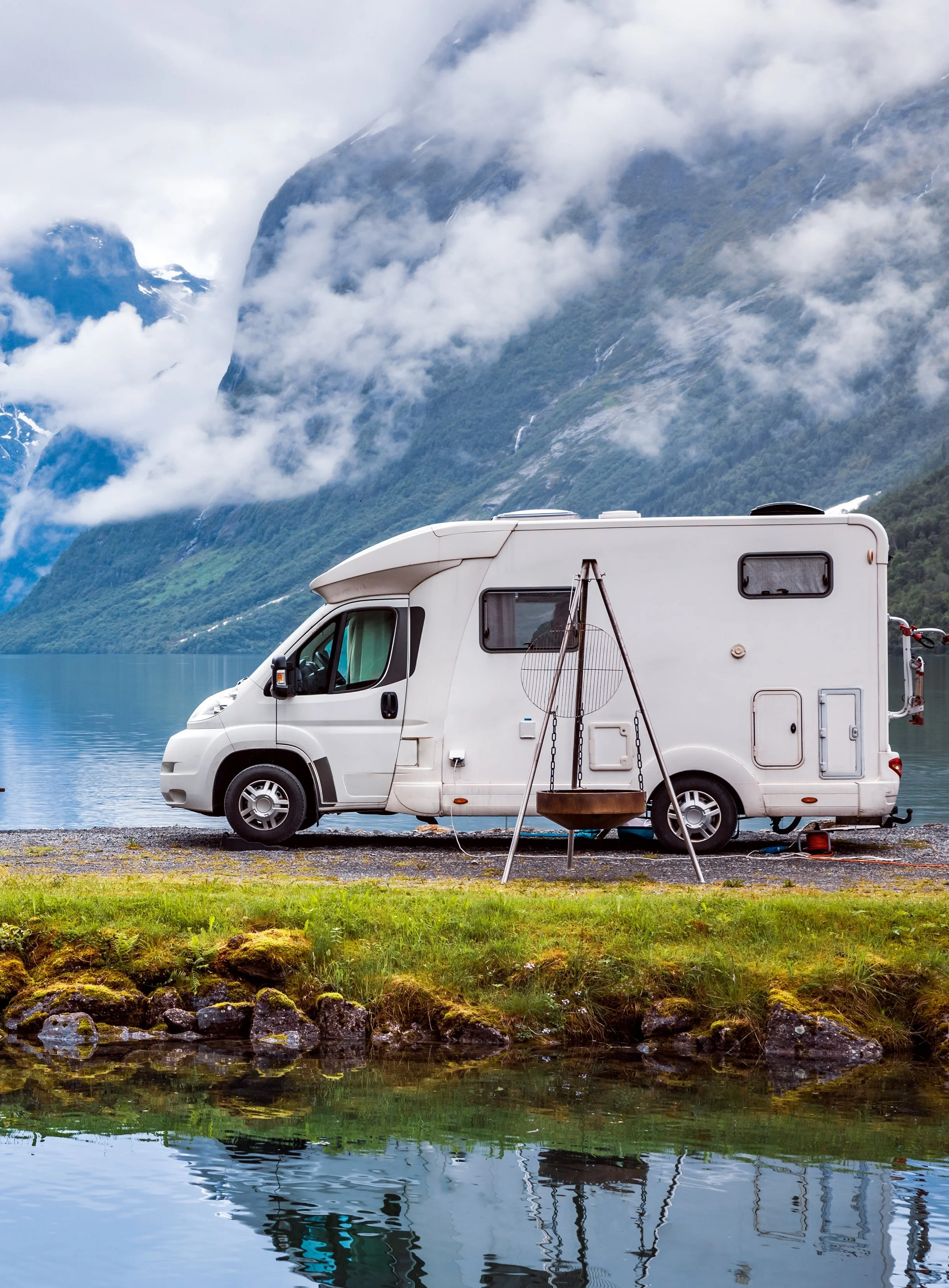 A white camper van parked near a lake with mountain scenery in the background, including clouds, water, and greenery.