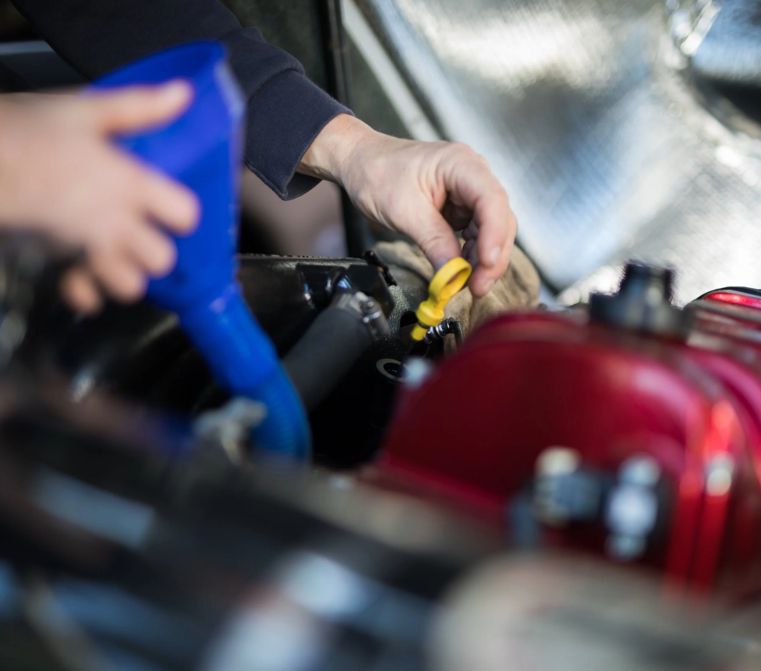 Close-up of a person working on a car engine, with their hand using a yellow tool and wearing a dark long sleeve shirt, with some blue and red wires visible in the background.