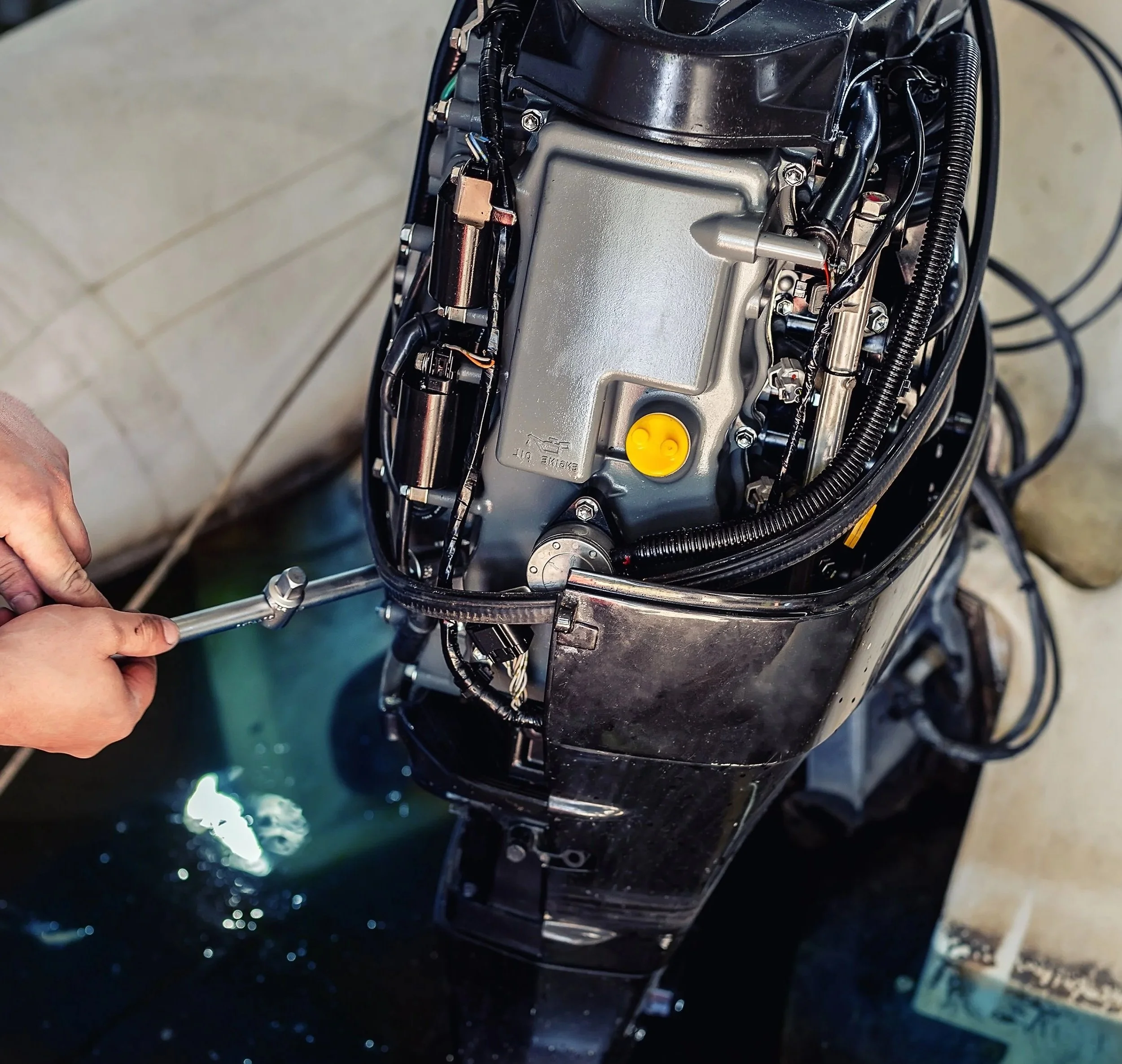 Close-up of an outboard motor showing wires, bolts, and mechanical parts being worked on with a hand holding a tool.