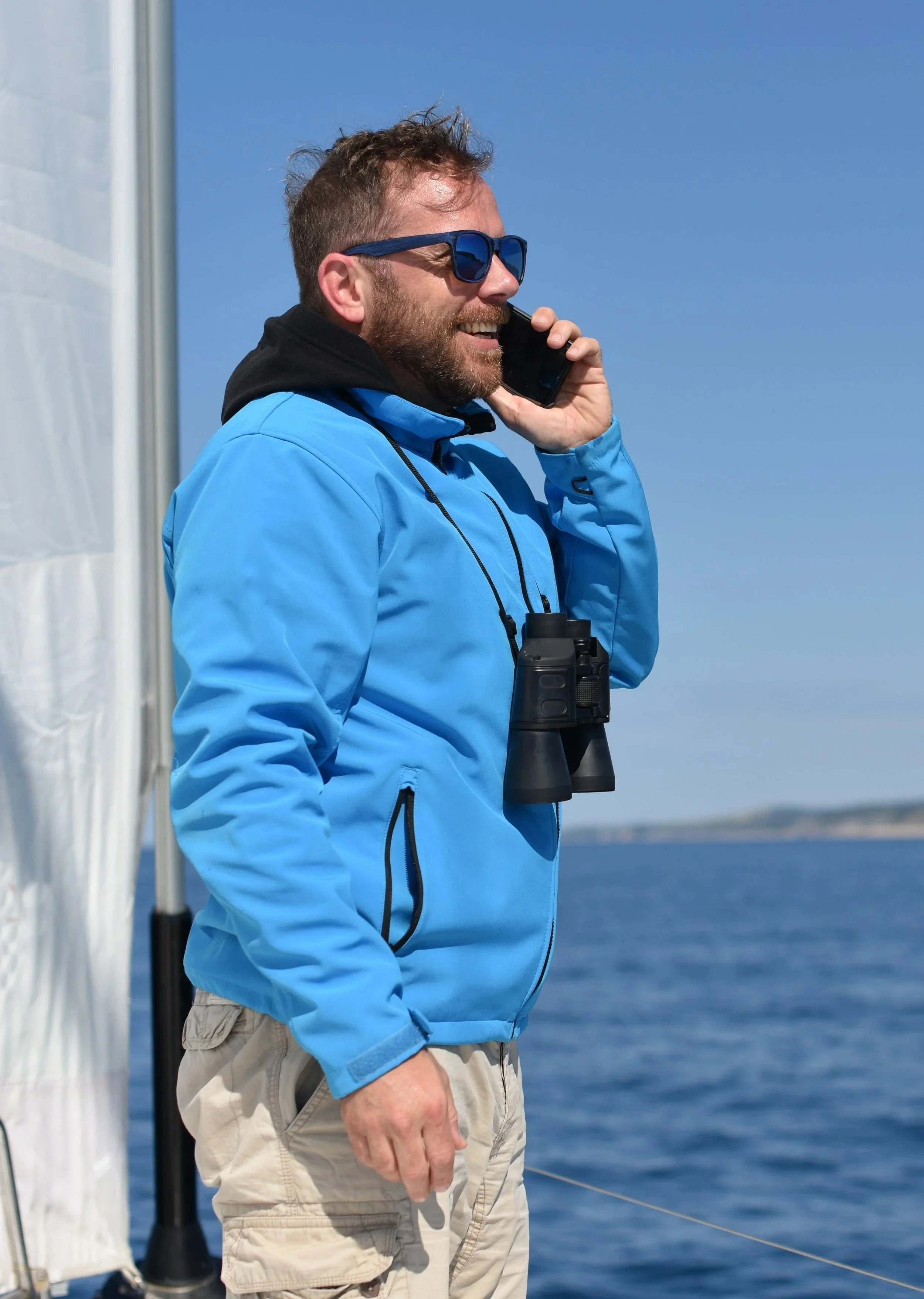Man with beard and sunglasses talking on a cell phone aboard a boat, wearing a blue jacket and beige pants, with ocean and clear blue sky in background.