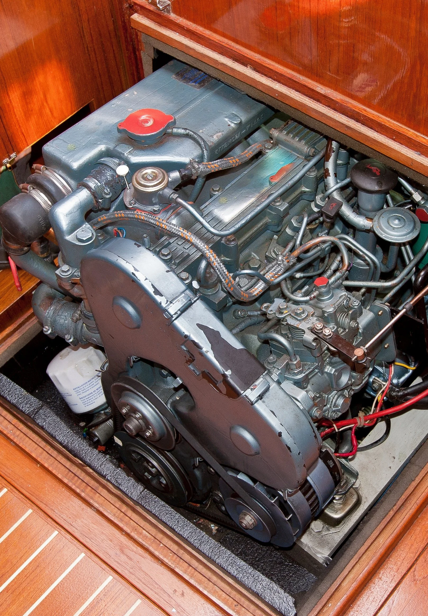 A marine engine inside a wooden enclosure with a wooden floor, showing various pipes, belts, and metallic parts.