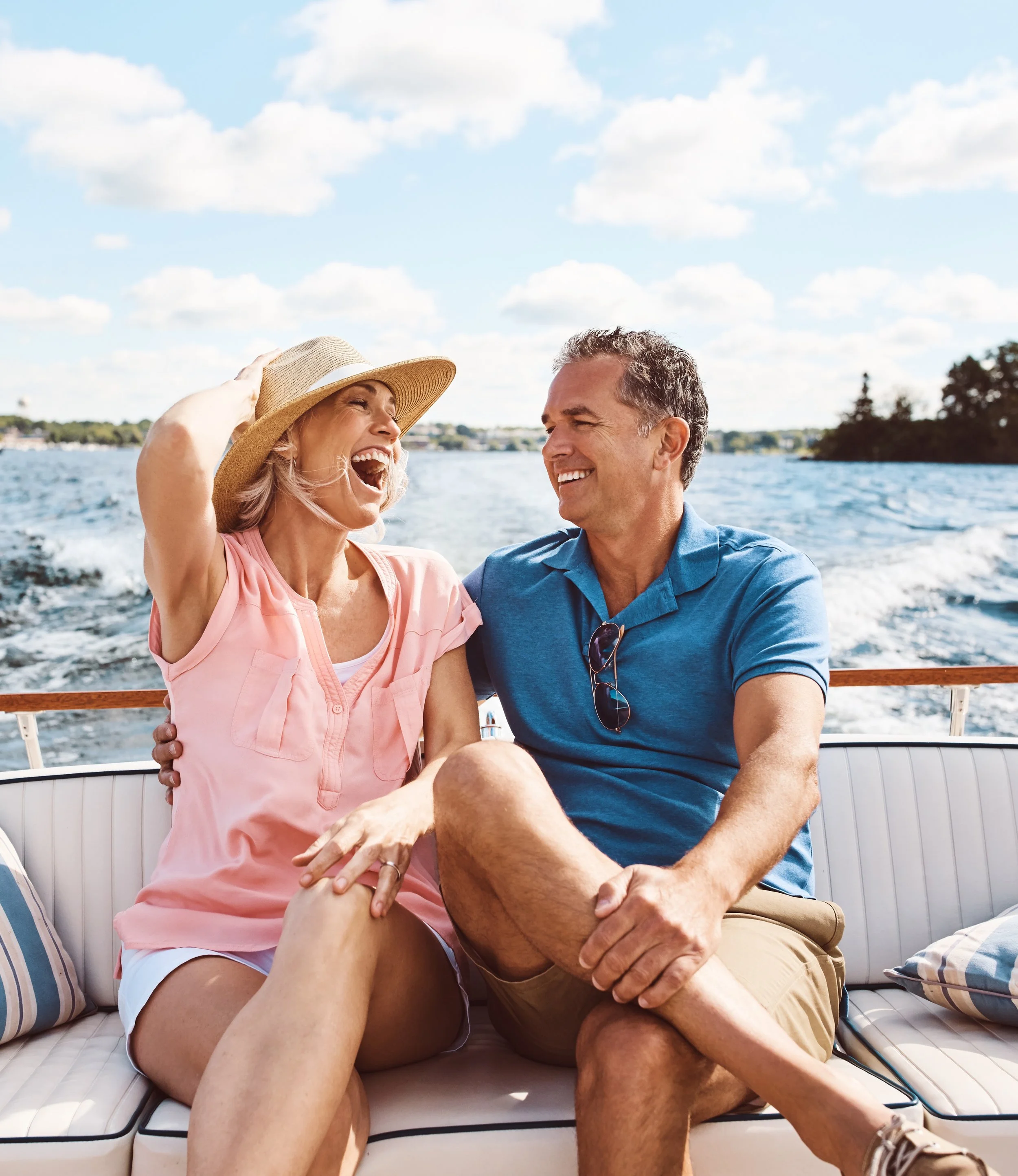 A joyful older woman with blonde hair wearing a wide-brimmed straw hat and a light pink shirt, laughing and holding her hat, is sitting on a boat with an older man who has gray hair and is wearing a blue shirt and beige shorts. They are outdoors on a sunny day with blue skies, scattered clouds, and water in the background.