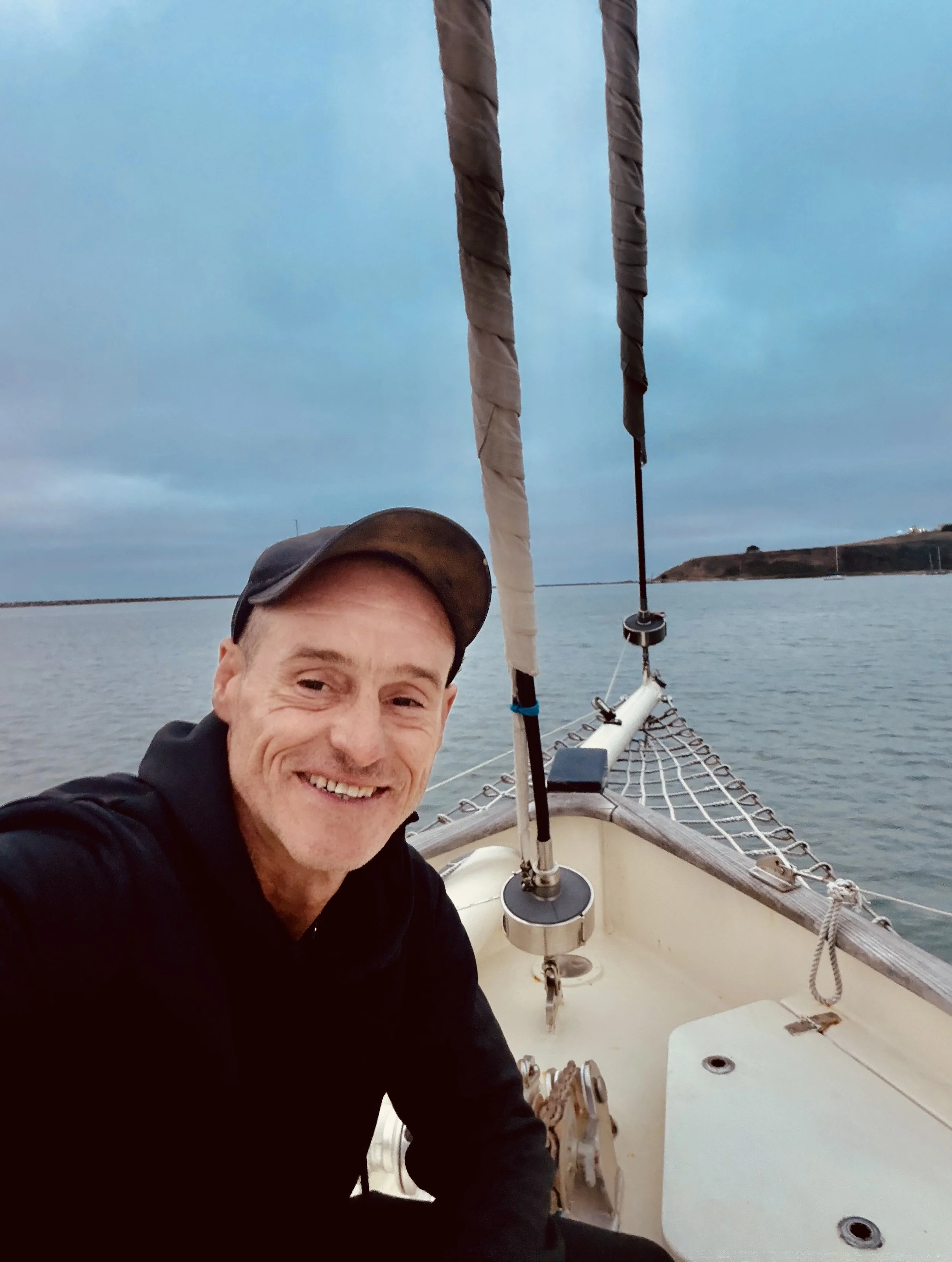 A man smiling on a sailboat at sunset or early evening, with the ocean and an island or coastline in the background, wearing a dark jacket and cap, with sailboat rigging and netting visible.
