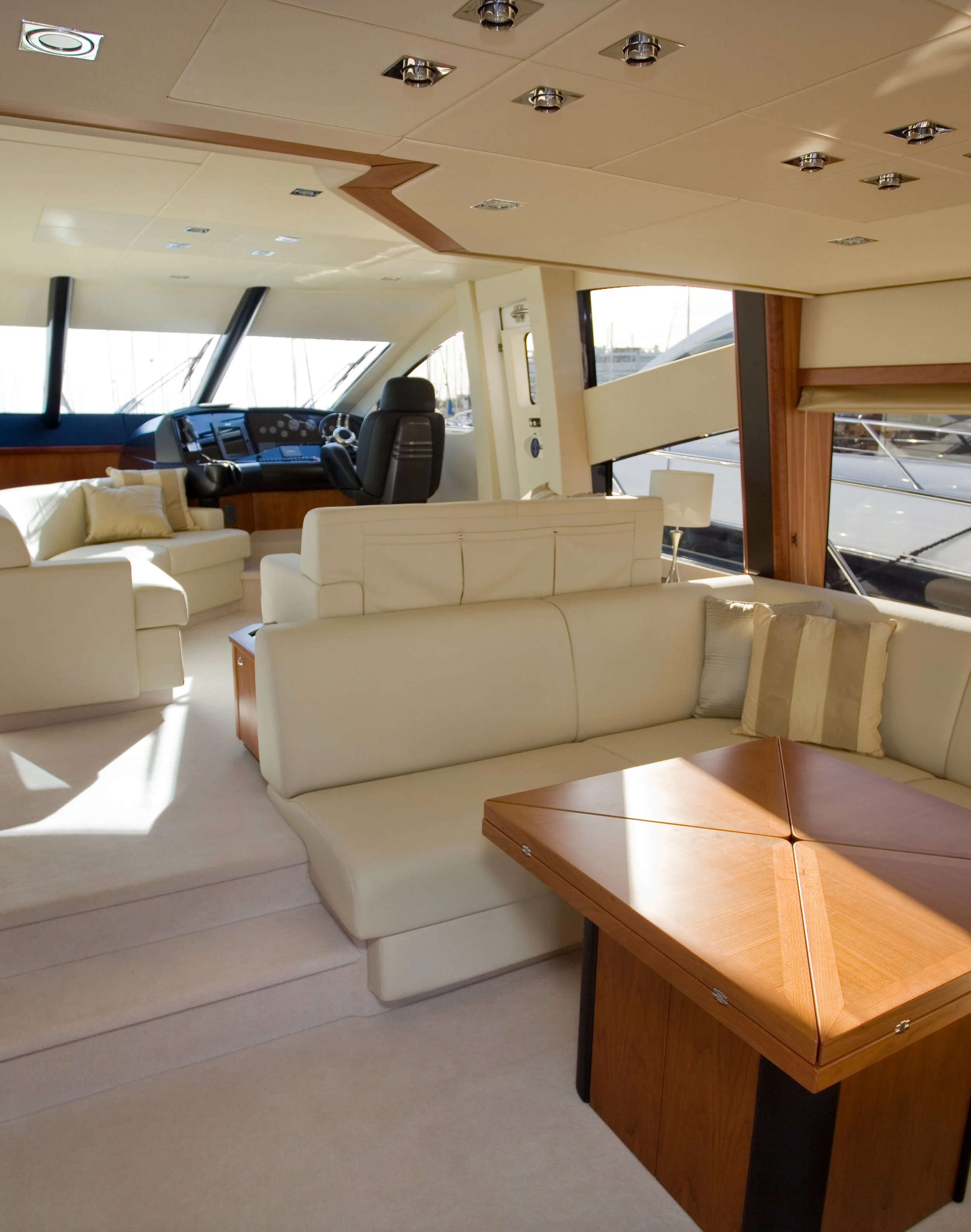 Interior of a yacht with cream-colored couches, a wooden table, and navigation controls in the helm area with windows showing a marina outside.