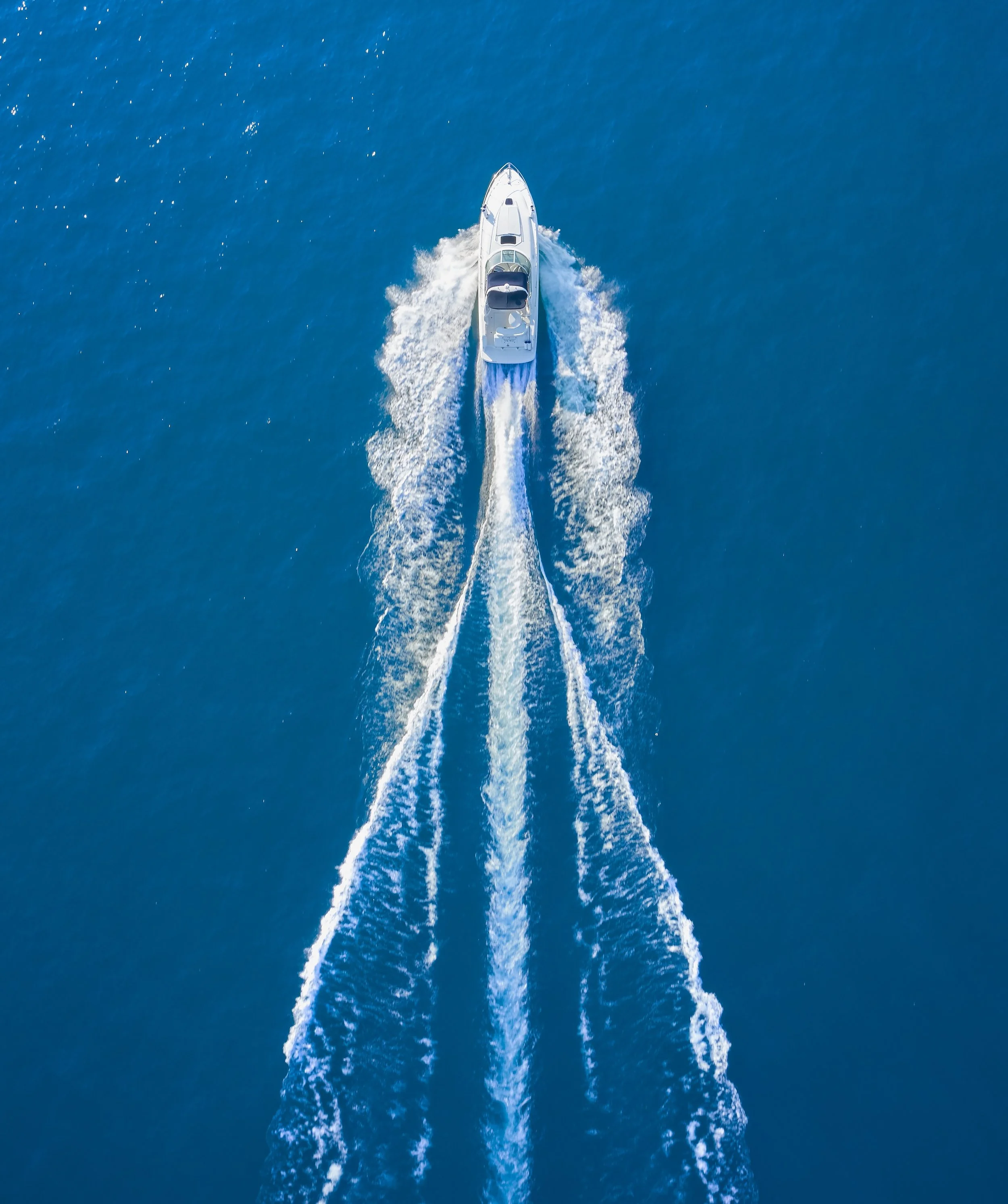 Top-down aerial view of a white yacht cruising through bright blue ocean water, leaving a foamy wake behind.