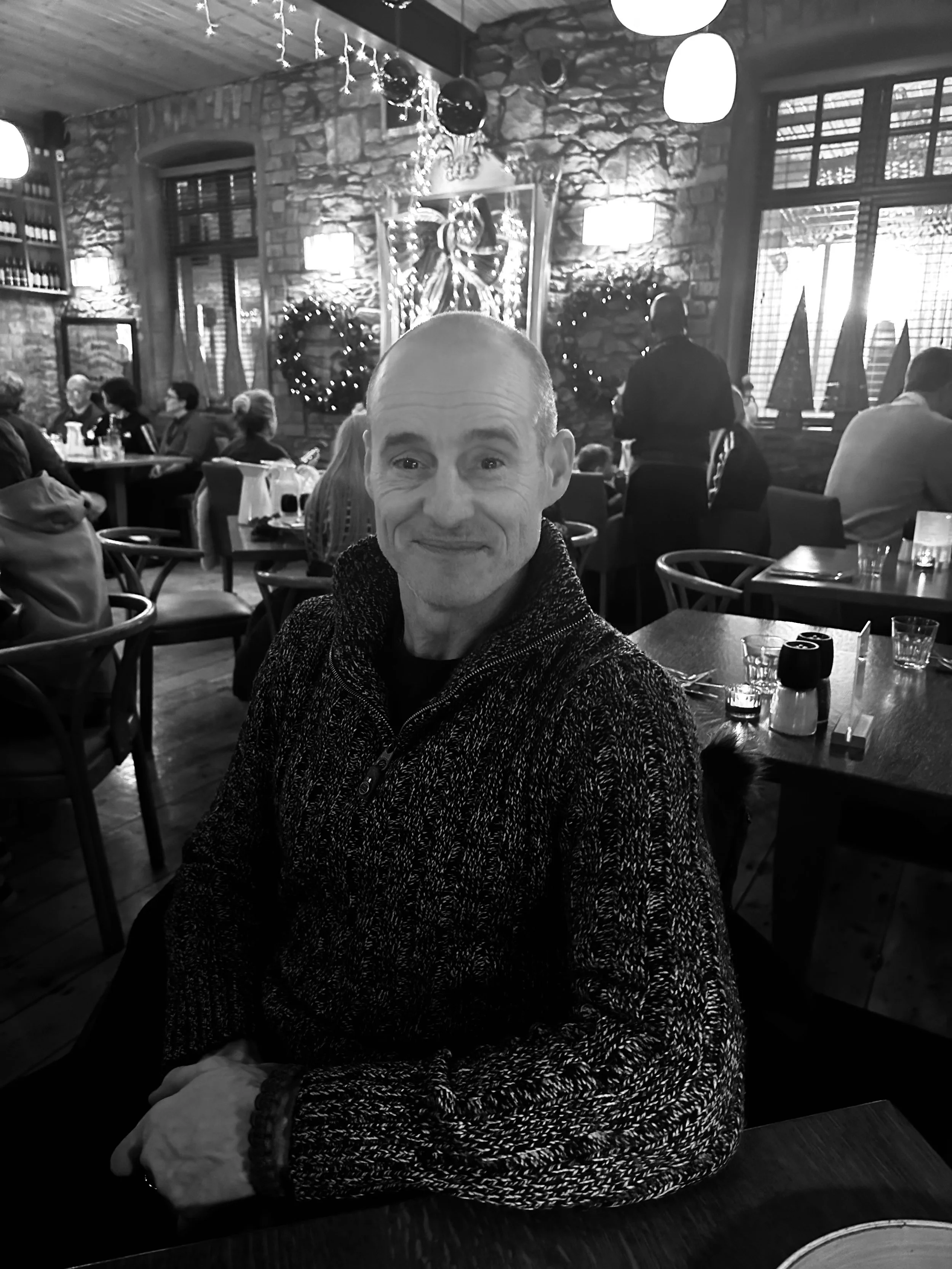 A man with a bald head smiling at the camera inside a cozy restaurant decorated for Christmas, with guests sitting at tables and festive wreaths and ornaments on the walls.
