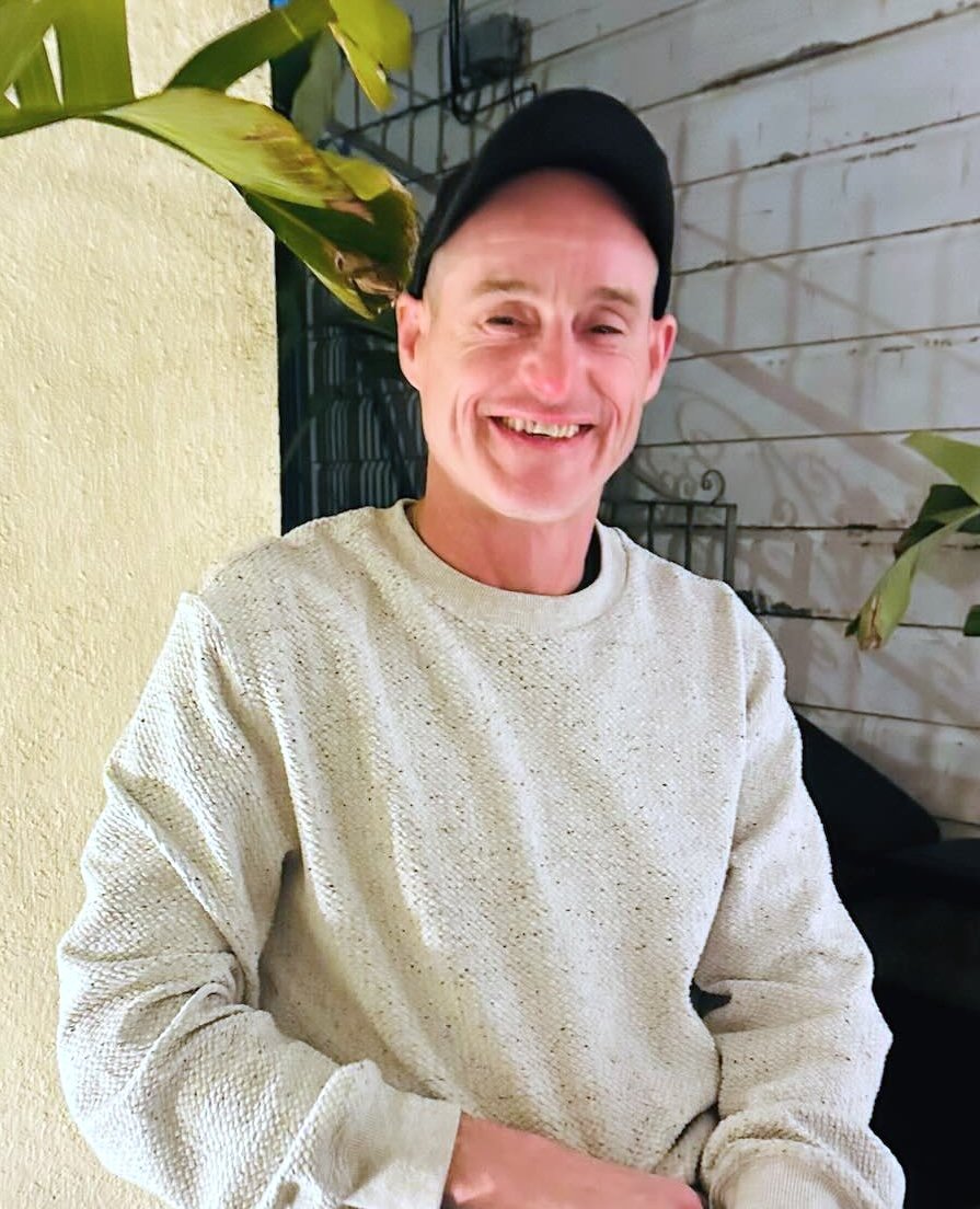 A smiling man wearing a black cap and a cream-colored sweater, sitting against a light yellow wall with plants around him.