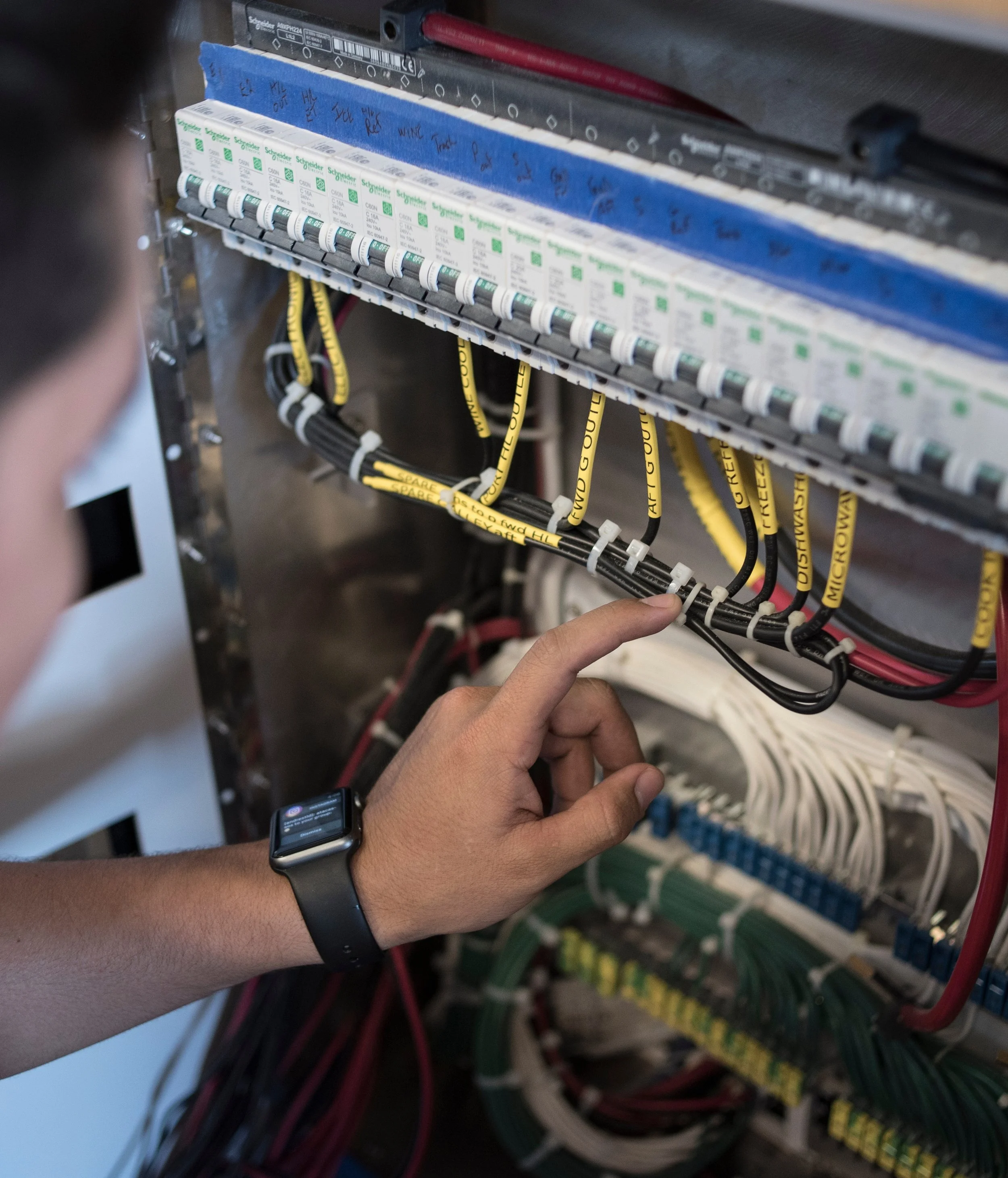 Close-up of a person's hand working on an electrical panel with white circuit breakers labeled with the brand 'Schneider.' The person is wearing a smartwatch.