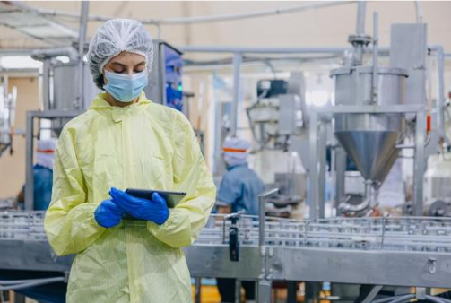 A person in protective clothing, including a yellow gown, blue gloves, a hair cover, and a face mask, standing in a manufacturing or processing facility, holding a tablet.