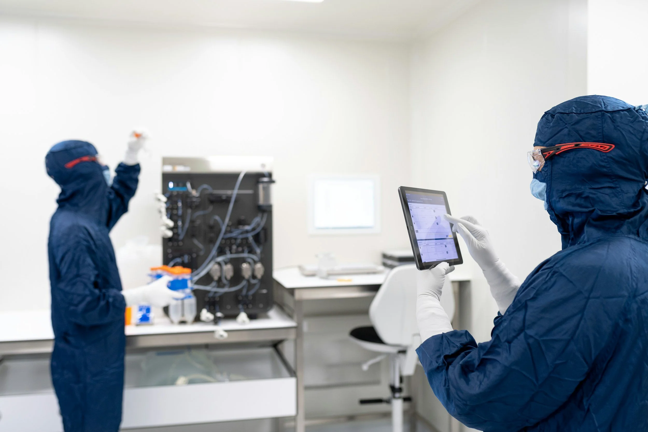 Two scientists in protective gear working in a laboratory with laboratory equipment and a tablet device.