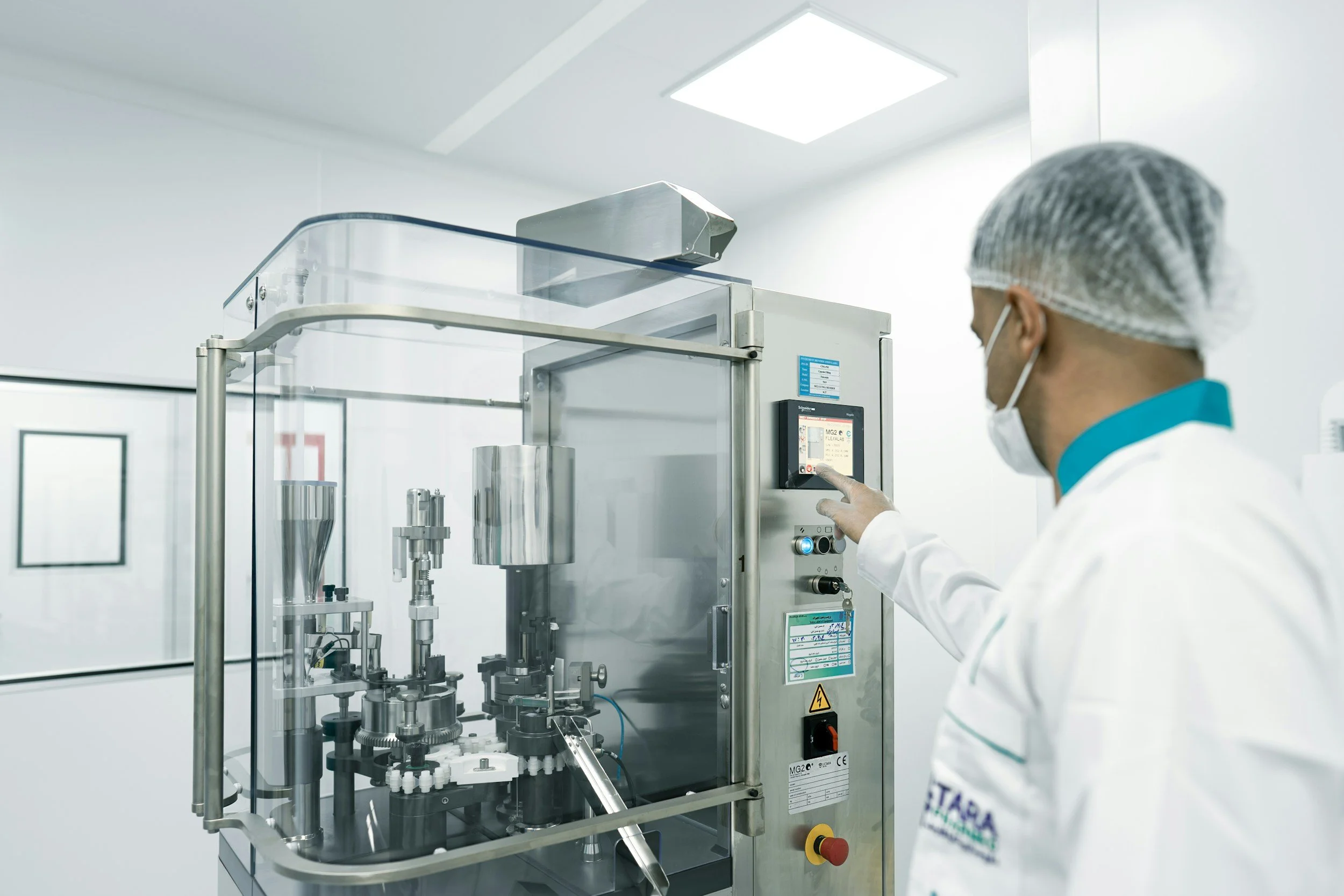 A scientist in a lab coat, hairnet, and mask operating a machine in a clean laboratory environment.