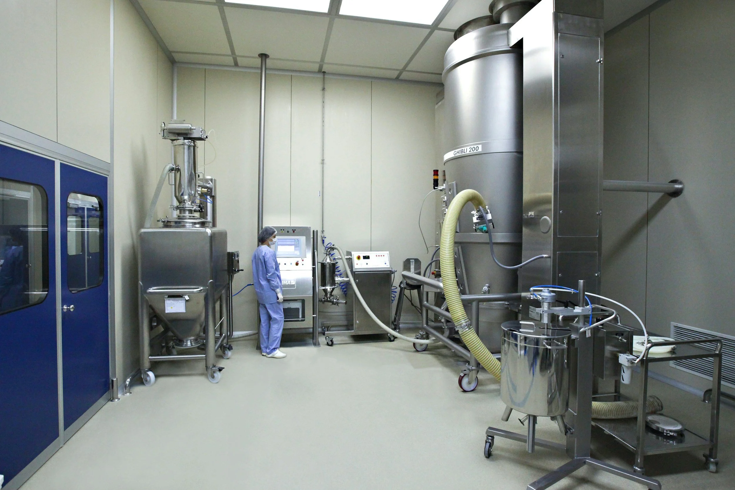 A scientist in scrubs and a hairnet working with laboratory equipment in a clean room. The room has stainless steel machinery, hoses, and a control panel.