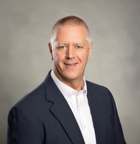Professional headshot of a middle-aged man with short gray hair, wearing a dark suit jacket and white dress shirt, smiling slightly against a neutral background.