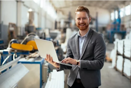 A man in a gray suit smiling while holding a laptop in an industrial or manufacturing setting.