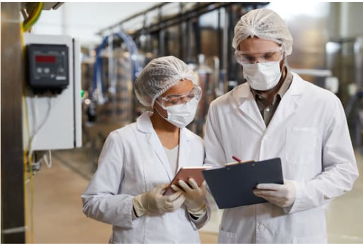 Two factory workers wearing white lab coats, hairnets, masks, and gloves inspecting documents in an industrial setting.
