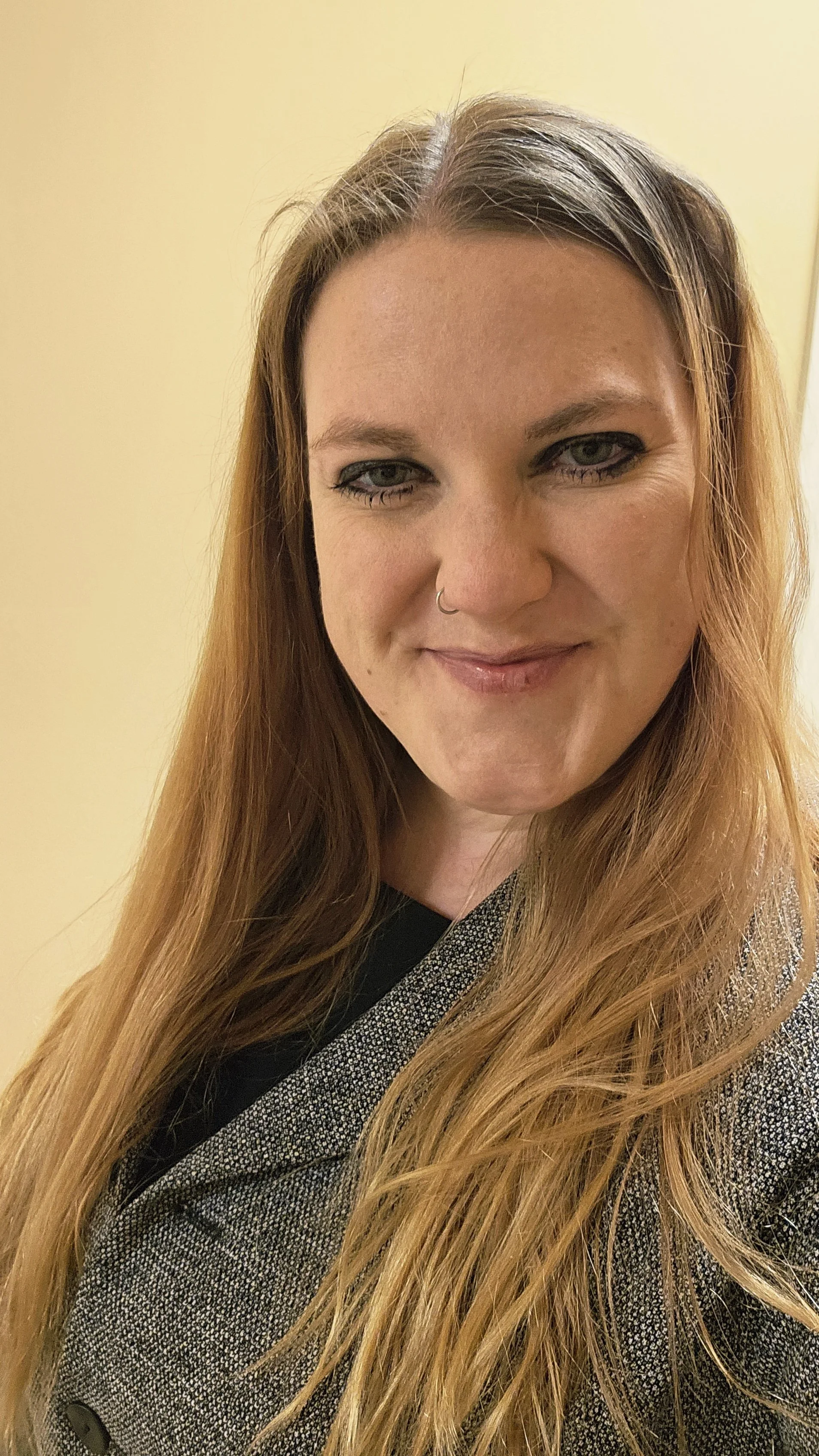 Close-up of a woman with long red hair, wearing a blue and white patterned blazer, against a beige background.