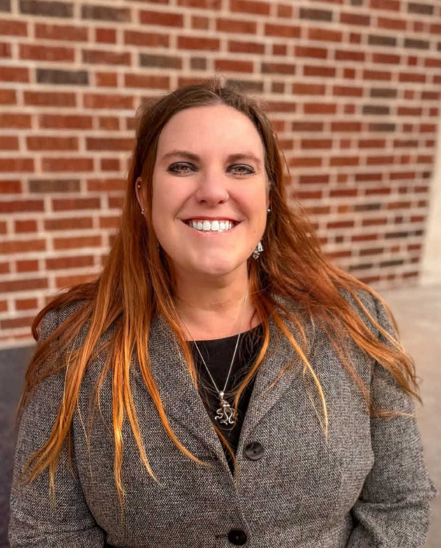 A woman with long reddish-brown hair smiling in front of a brick wall.