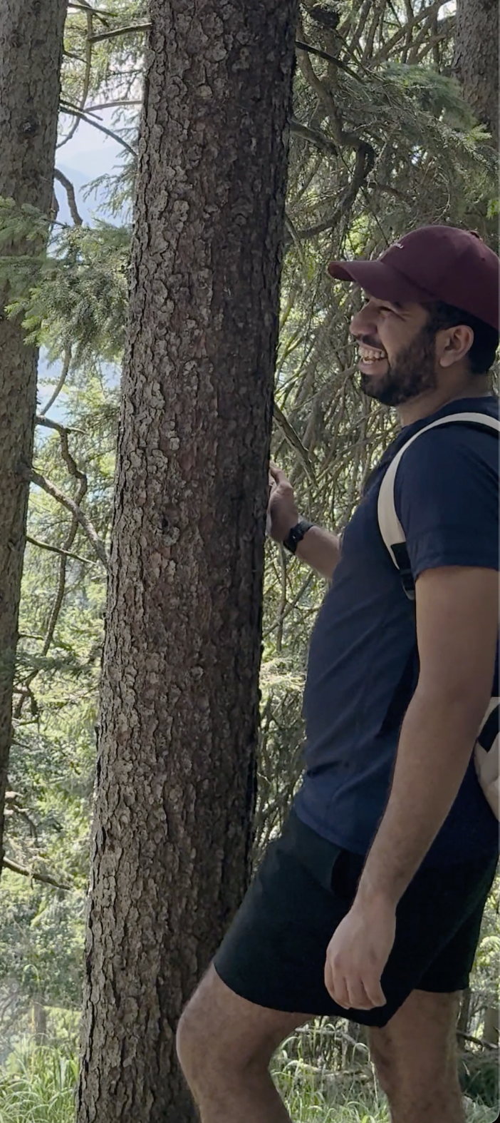 A man in a maroon cap and navy T-shirt smiling cheerfully as he leans on a tree trunk in a forest, with green foliage and trees in the background.