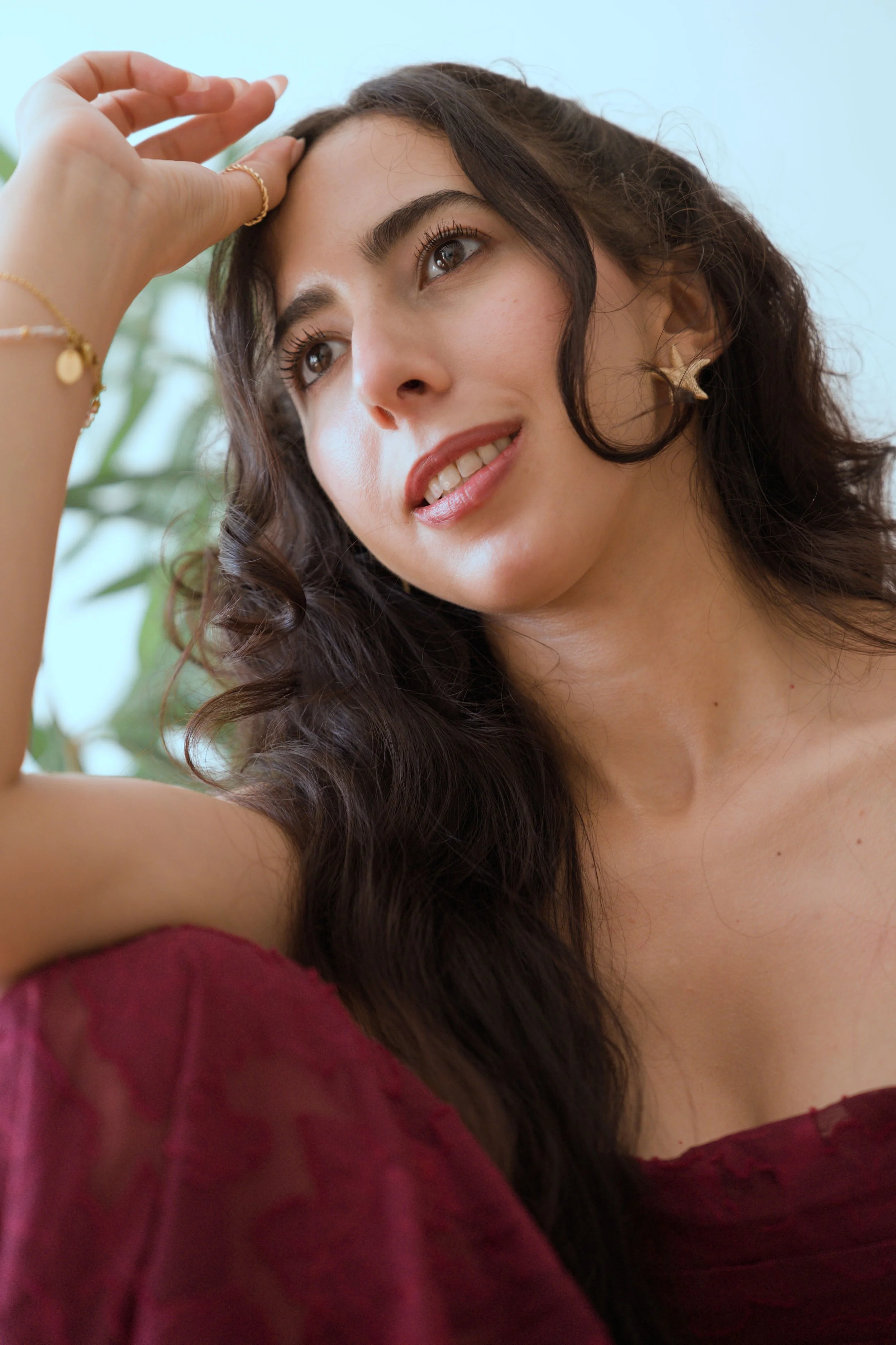 Ihssane with long, wavy brown hair smiling and looking away, wearing gold jewelry and a burgundy off-the-shoulder top, with a blurred green plant in the background.