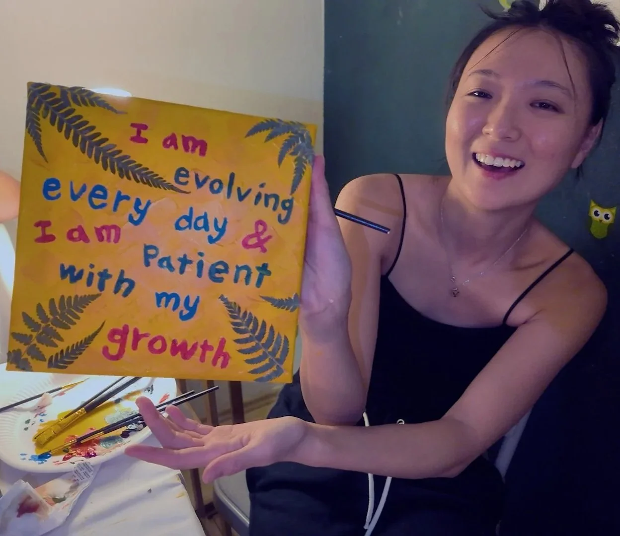 A smiling woman holding a colorful canvas with the words "I am evolving every day & I am patient with my growth," surrounded by painted fern leaves. She is wearing a black spaghetti strap top and necklace, and she appears to be in an art studio or a creative space.