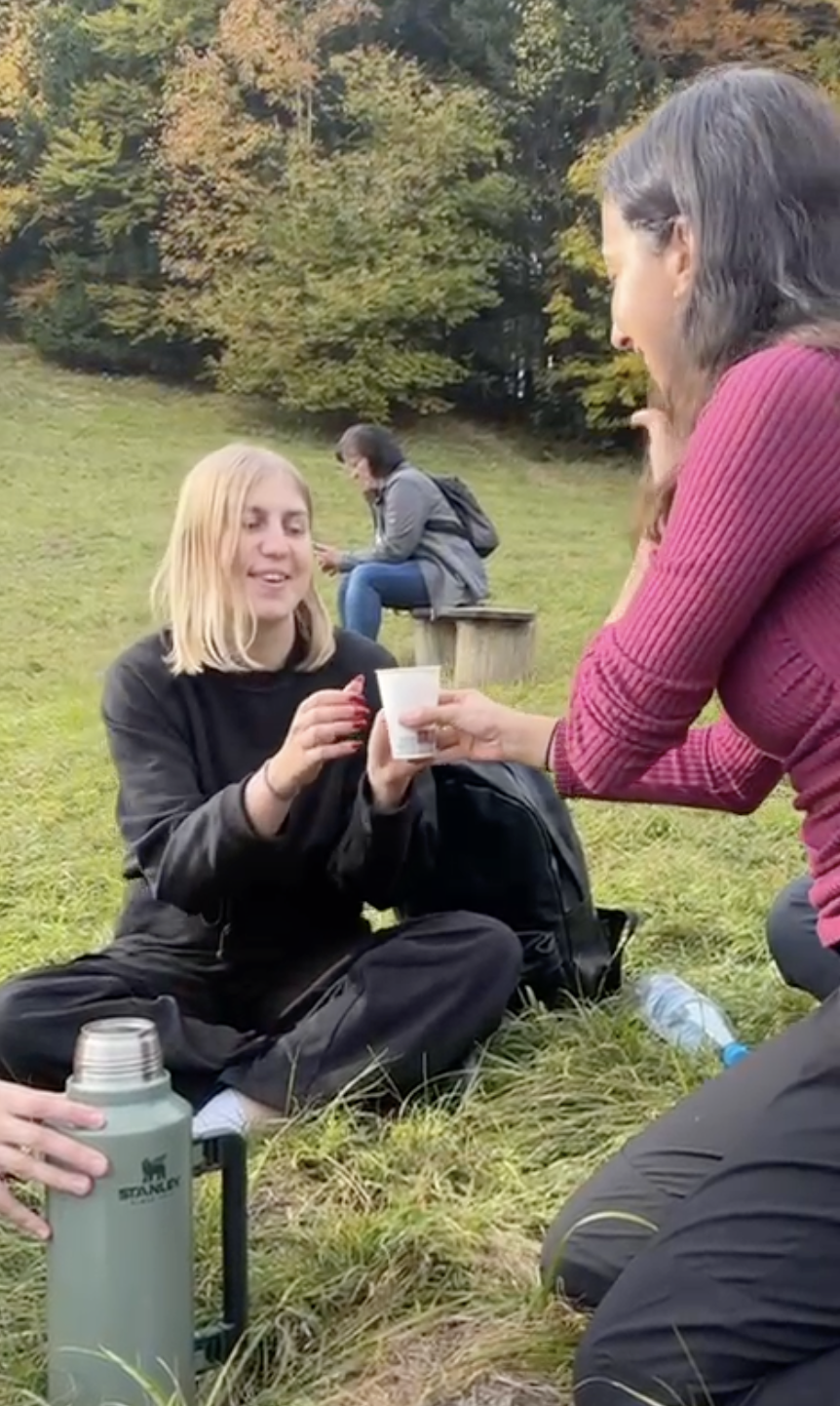 Two women outdoors, one sitting on the grass and the other kneeling, exchanging a small cup, with a woman in the background sitting on a wooden bench using a phone.