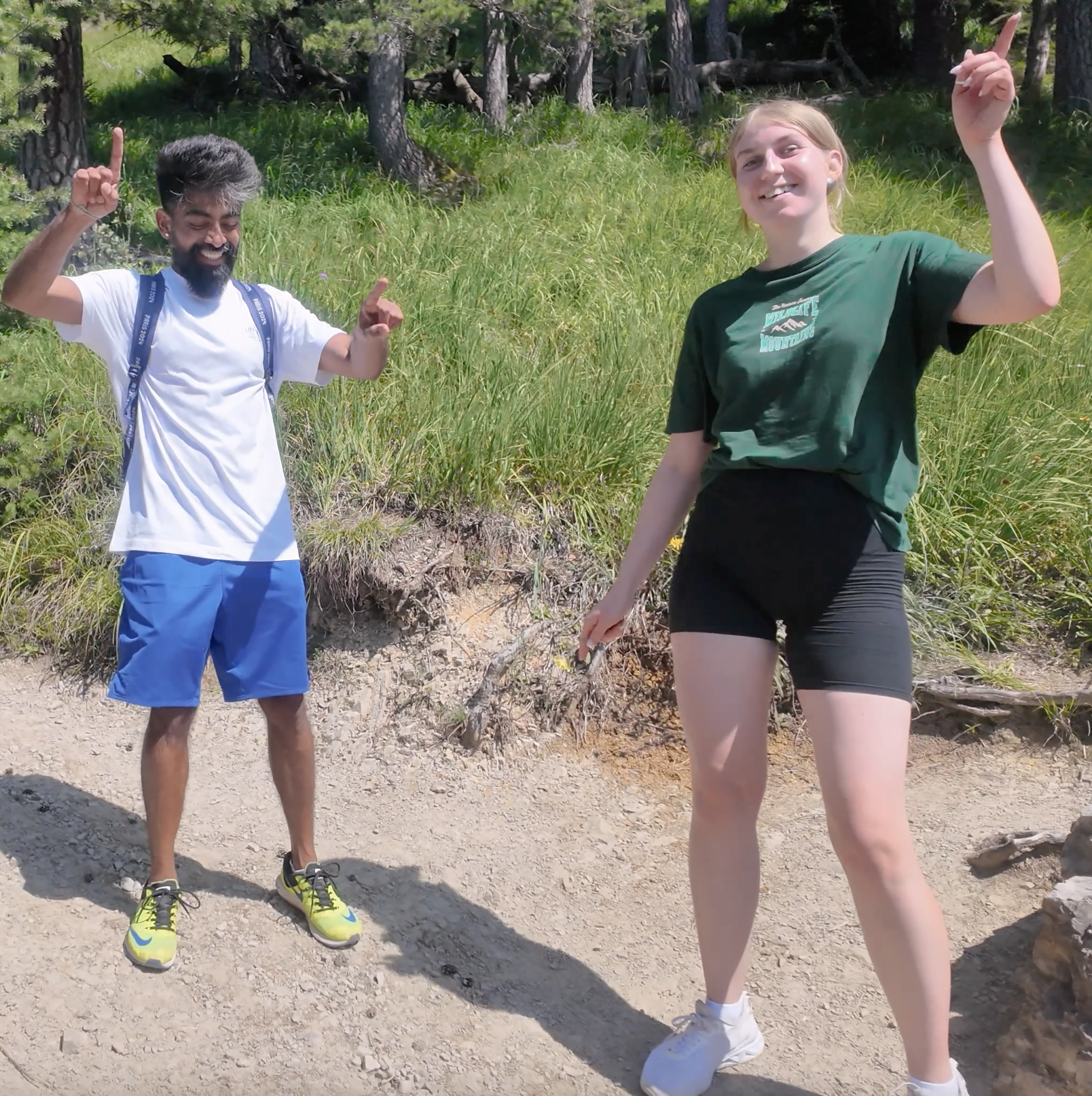 Two smiling young people, a man and a woman, standing outdoors on a dirt path with greenery and trees in the background. They are gesturing upward with one finger.